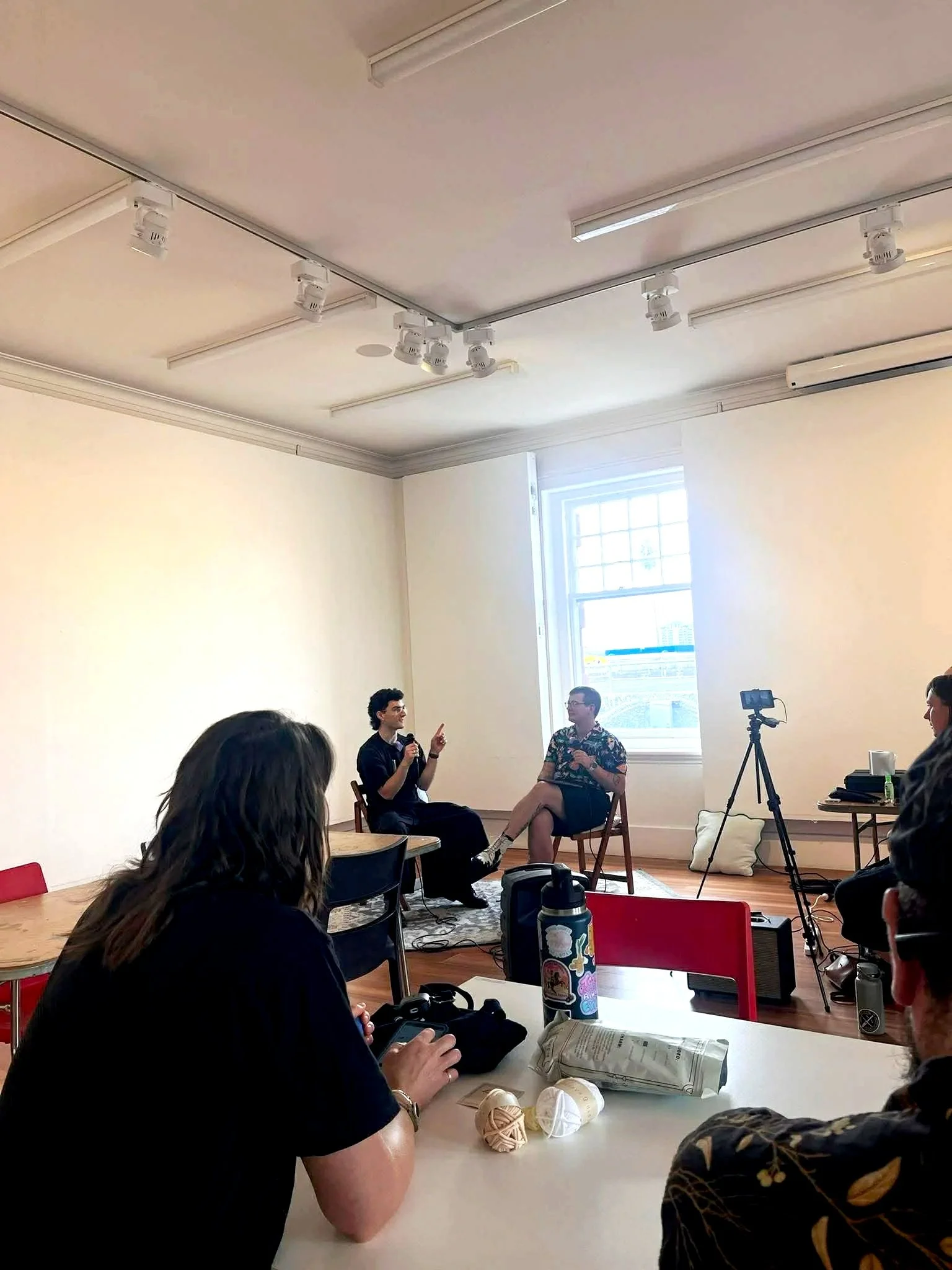 A discussion or interview taking place in a room with a person speaking into a microphone and another listening. Several people are seated around tables with items such as books, papers, and yarn. A camera on a tripod is set up in the background.