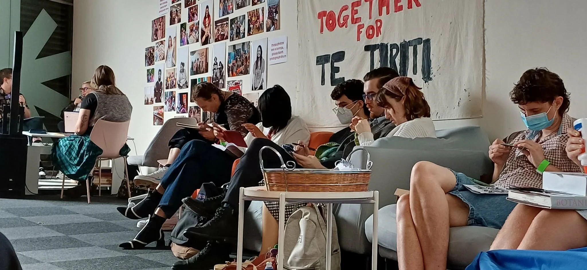 People sitting in a waiting area, some using smartphones and wearing masks, with a wall displaying photos and a large banner with the words 'Together for TE TRUI'.