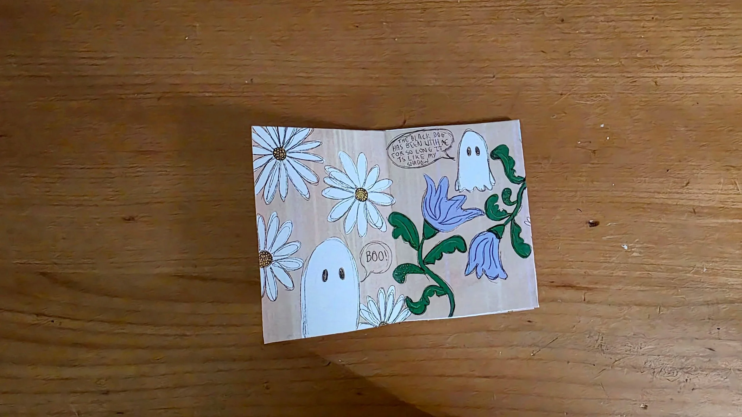 A hand-drawn comic on a piece of paper featuring white ghosts, flowers, and speech bubbles with text about a black dog. The background is a wooden surface.