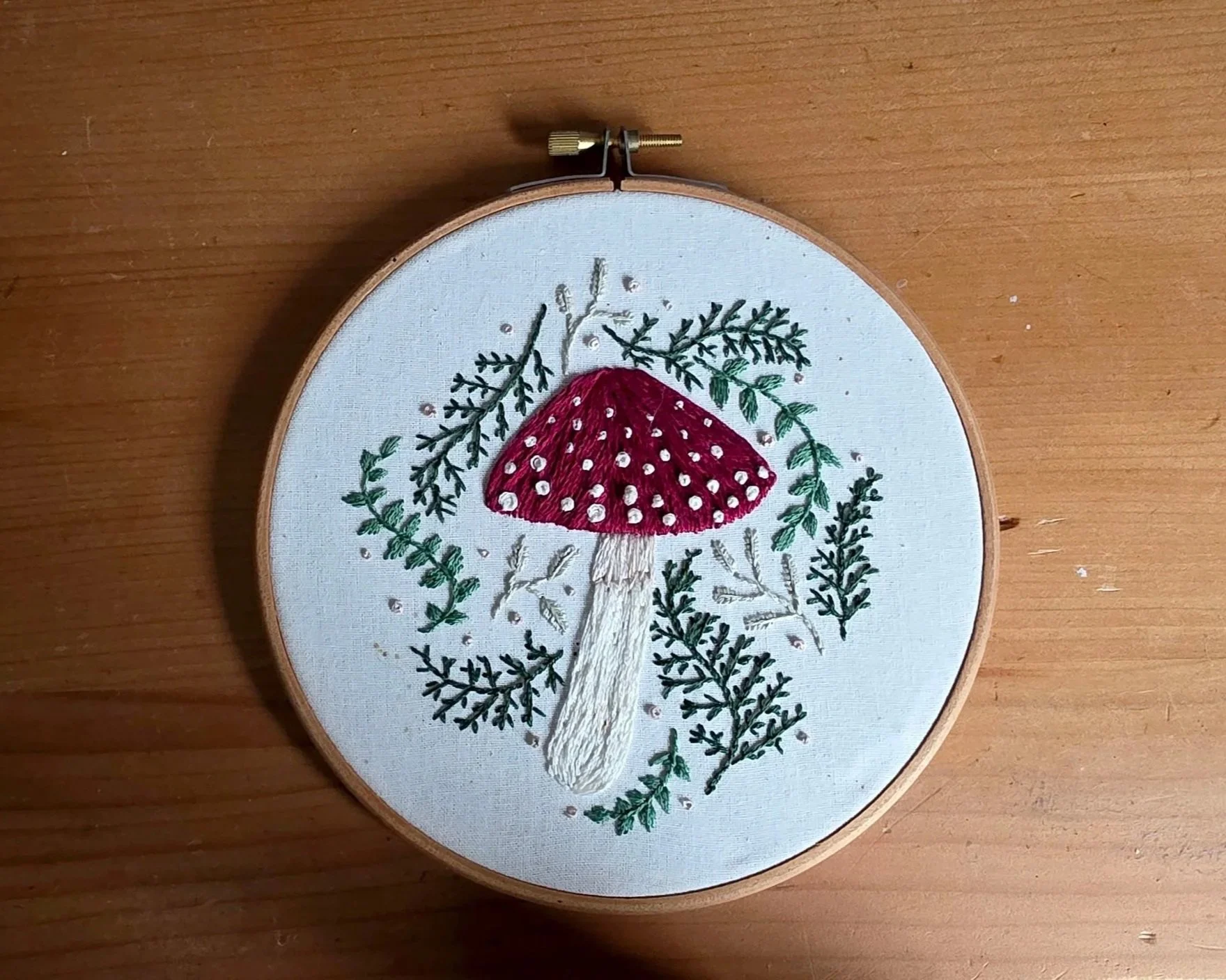 Embroidery of a red mushroom with white spots, surrounded by green foliage, on white fabric placed in an embroidery hoop.