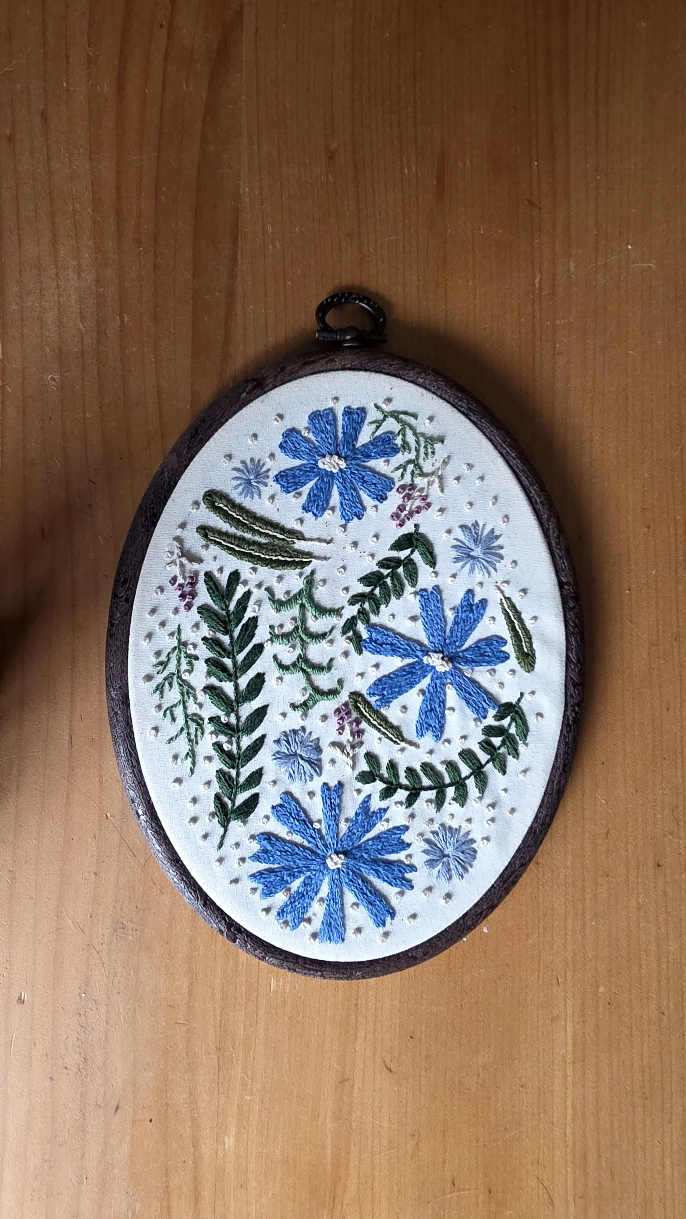 Embroidery hoop with hand-stitched blue flowers and green leaves on white fabric, on a wooden surface.