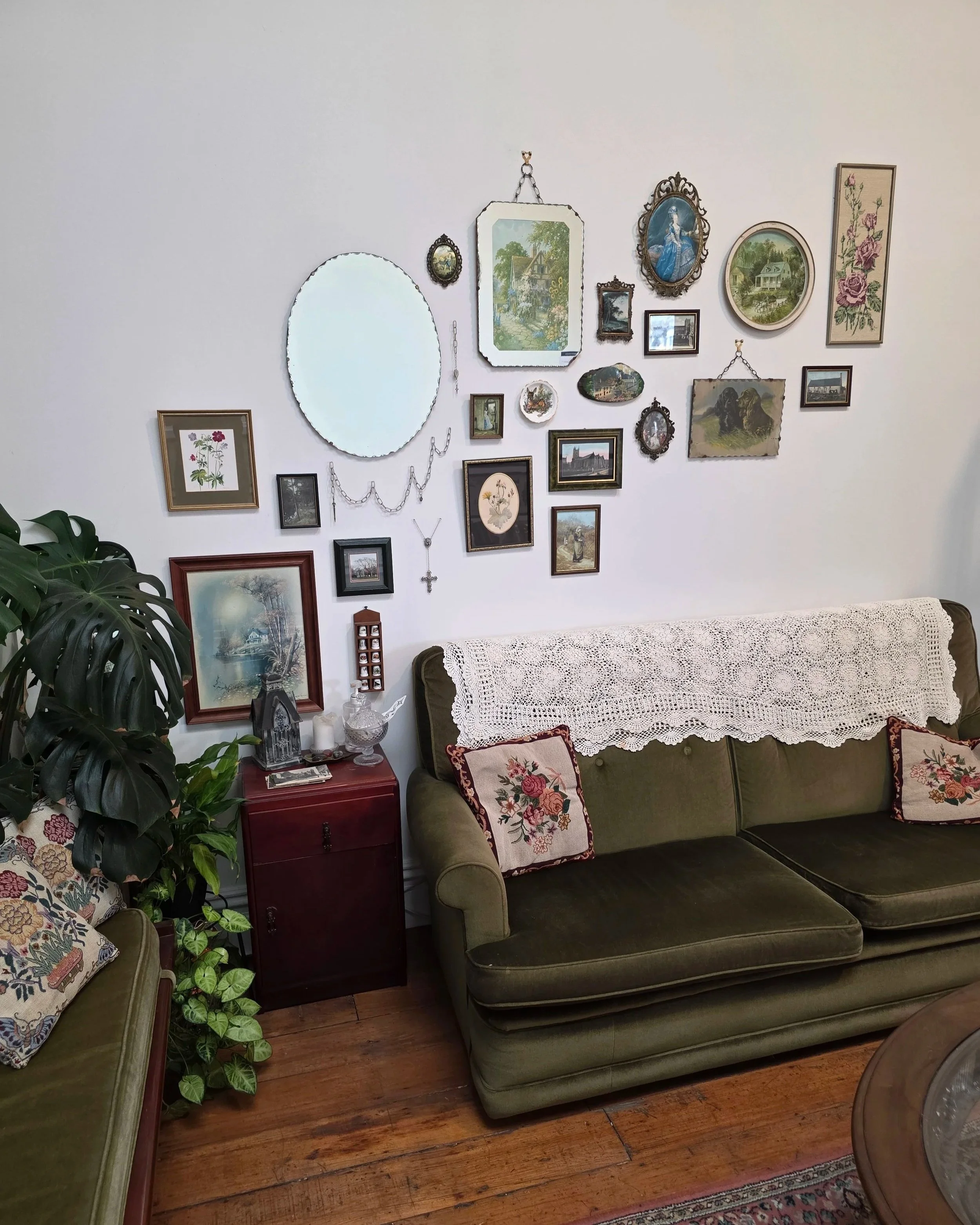 A vintage living room with green velvet sofa, floral cushions, a crocheted lace throw, antique frames and paintings on the wall, a small red side table with decorative items, and a large leafy plant. The floor is wooden with a decorative rug partially visible.