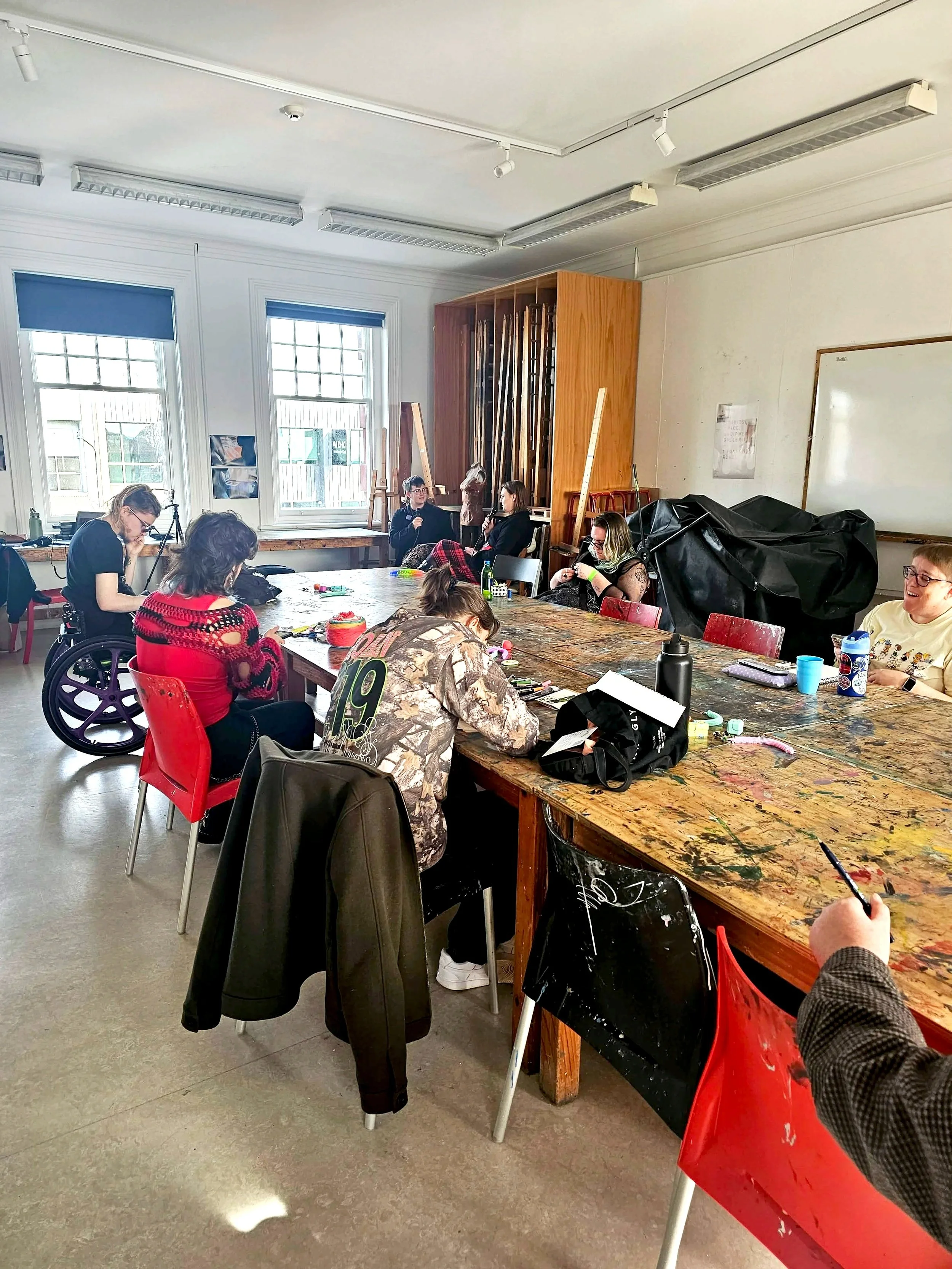 People sitting around a large, paint-stained wooden table in a bright room with white walls and large windows, engaged in conversation or working on art projects. There are various objects on the table, including water bottles, cups, and art supplies.
