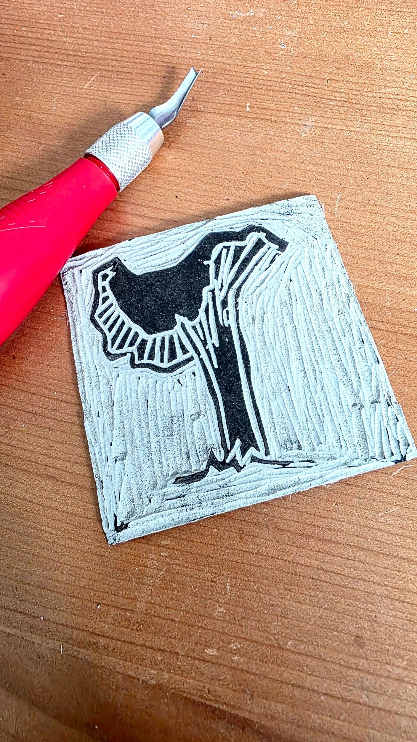 A linocut print of a wolf howling at the moon on a textured square piece of linoleum, with a carving tool and a piece of paper nearby, all on a wooden surface.