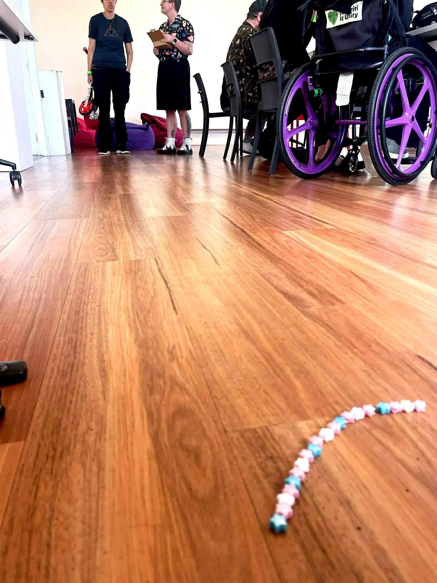 View of a room with a wooden floor, showing some people in the background and a wheelchair with purple wheels on the right side. There are some pastel-colored candies on the floor in the shape of a heart in the foreground.
