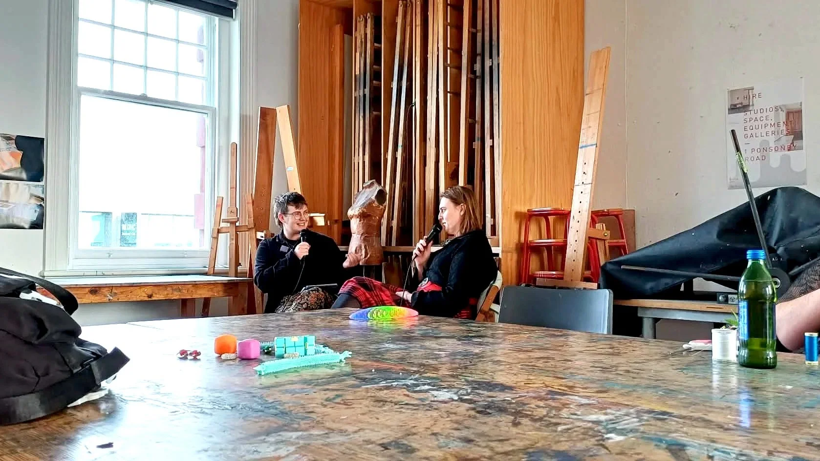 Two women sit at a table with microphones, engaging in conversation. The background features wooden paneling, art supplies, and a window letting in natural light.