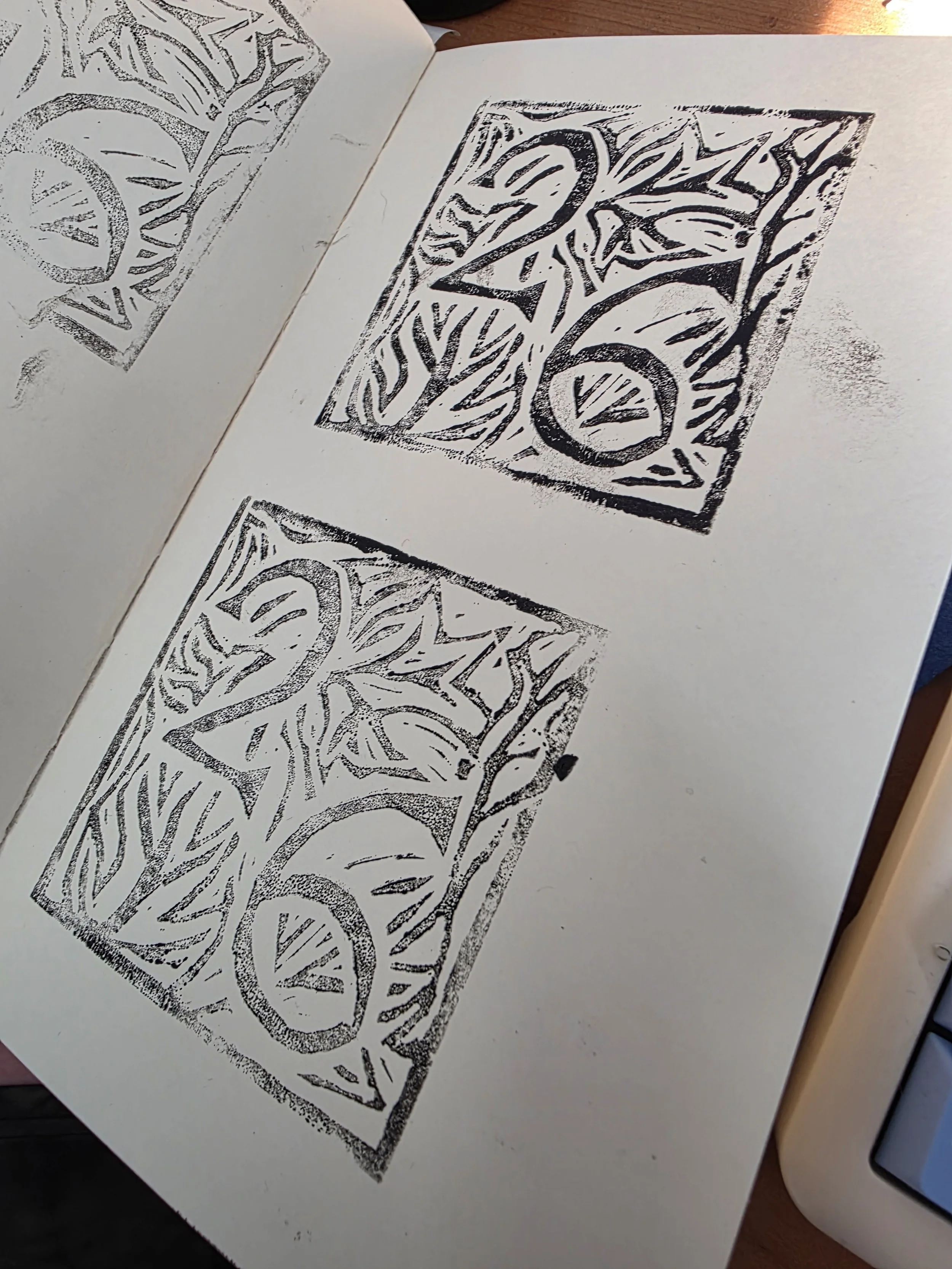 Black and white linocut print featuring a square with intricate, stylized leaves and circular shapes, shown in a sketchbook on a wooden surface.