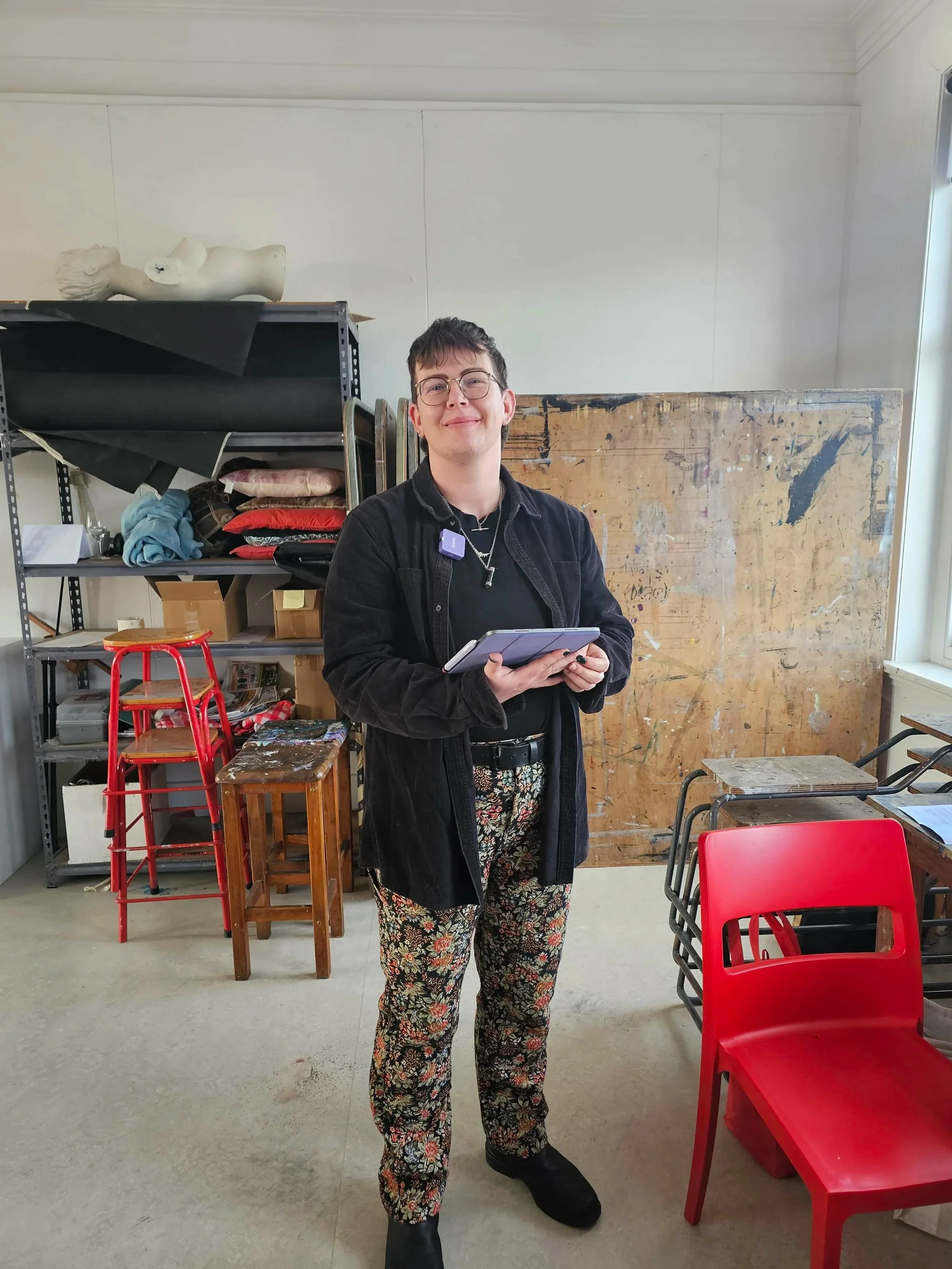 A person standing indoors holding a tablet, wearing glasses, a black jacket, floral pants, and boots, with shelves, chairs, and an easel in the background.