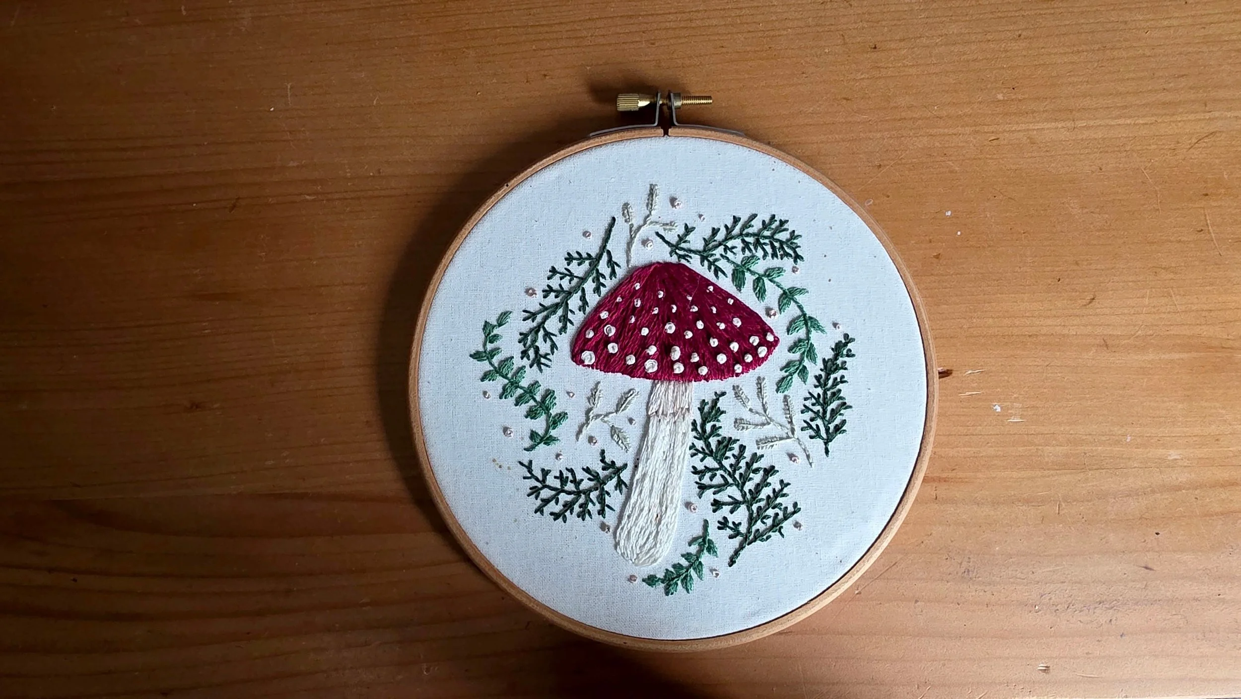 Embroidery hoop with a textile featuring a appliqué of a red mushroom with white spots, surrounded by embroidered green foliage and branches.
