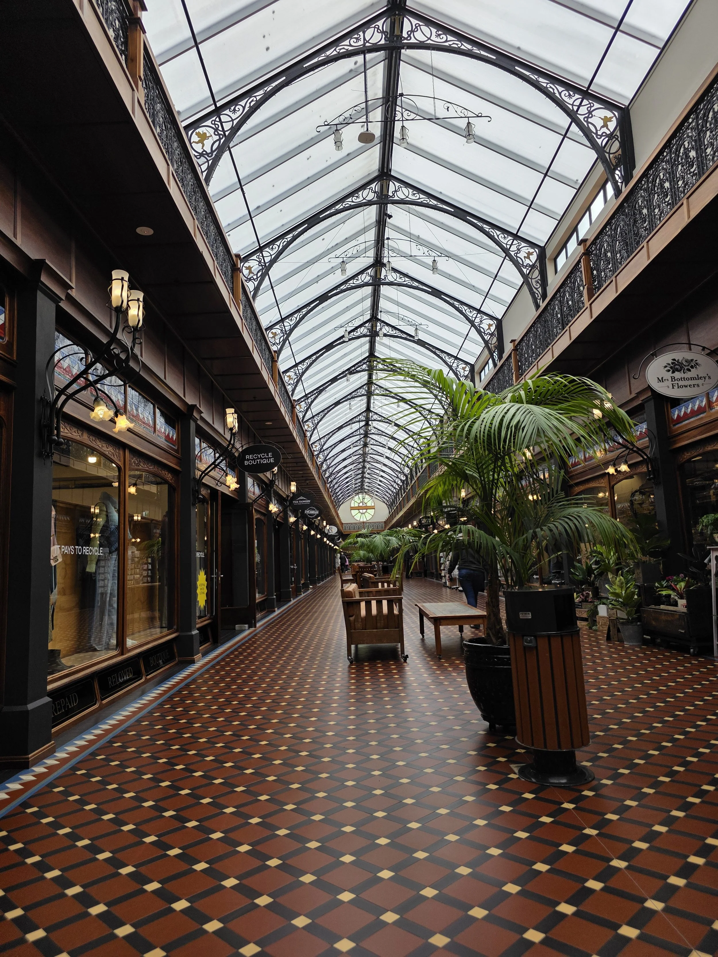 Indoor shopping gallery with decorative ironwork on the balconies, a glass roof, potted plants, and benches, in a historic style.