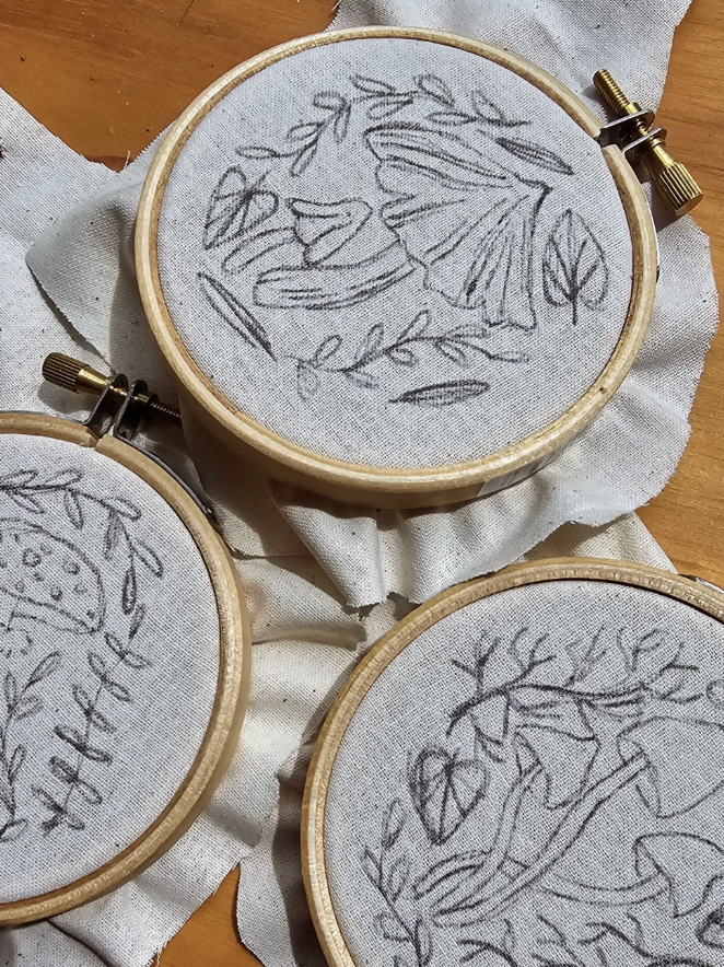 Embroidery hoops with fabric featuring pencil sketches of floral and nature-inspired designs, placed on a wooden surface