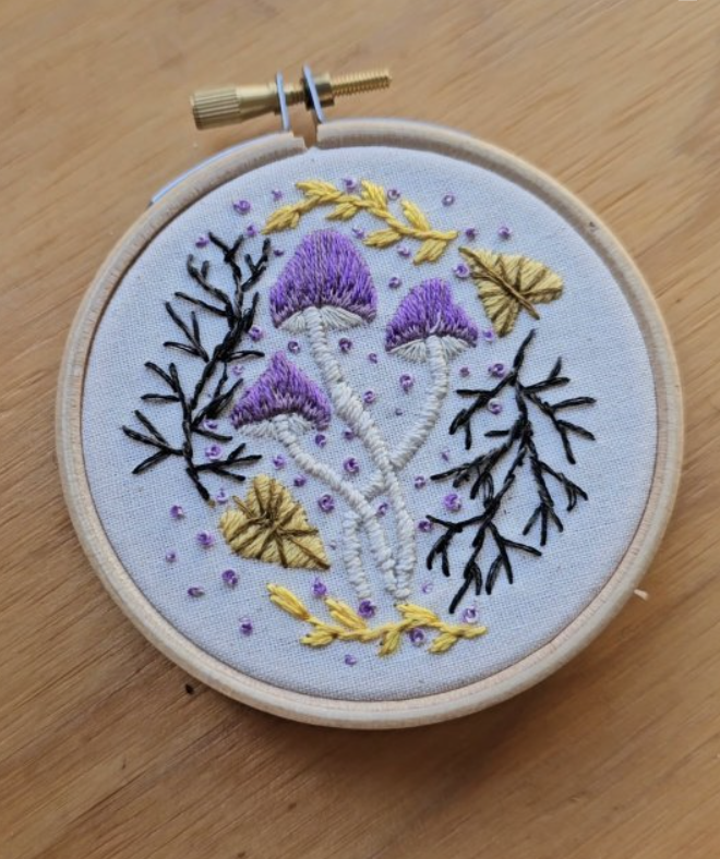 Embroidery hoop with stitched purple mushrooms, yellow leaves, black and yellow branches, and purple dots on white fabric.