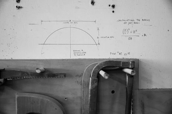 Part of a whiteboard with a diagram and formulas for calculating the radius of an arc. Below the whiteboard, there are metal equipment and a wooden chair in a workshop or classroom setting.