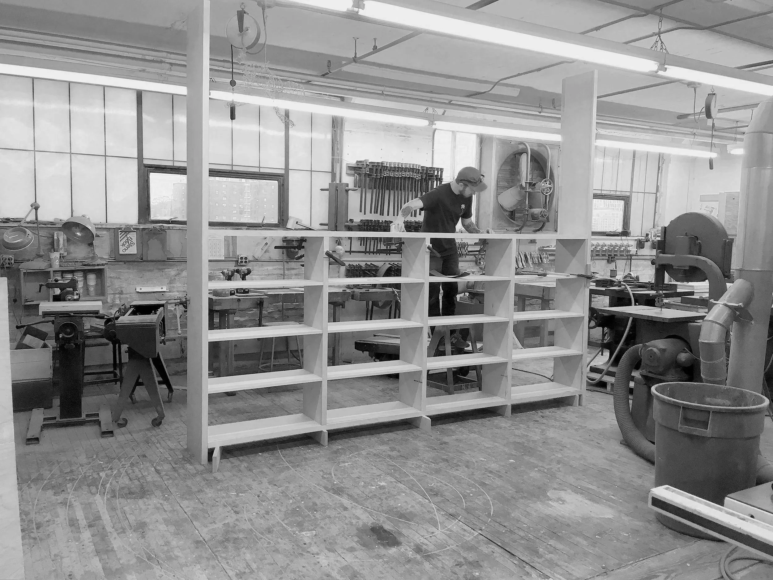 A black and white photo of a woodworking shop with a man working on a wooden frame, surrounded by various tools, equipment, and storage shelves.