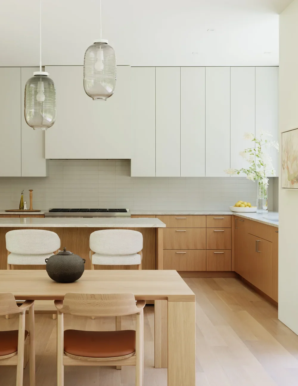 A modern kitchen with white upper cabinets and wooden lower cabinets, a white countertop, two pendant lights, a dining table with four wooden chairs, and a vase with white flowers on the countertop.