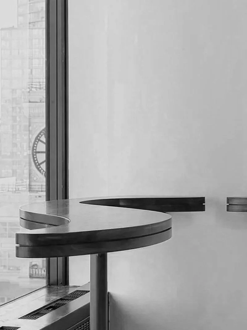 A minimalist modern interior with a curved wooden table, a window showing a clock in a cityscape background, and plain walls.