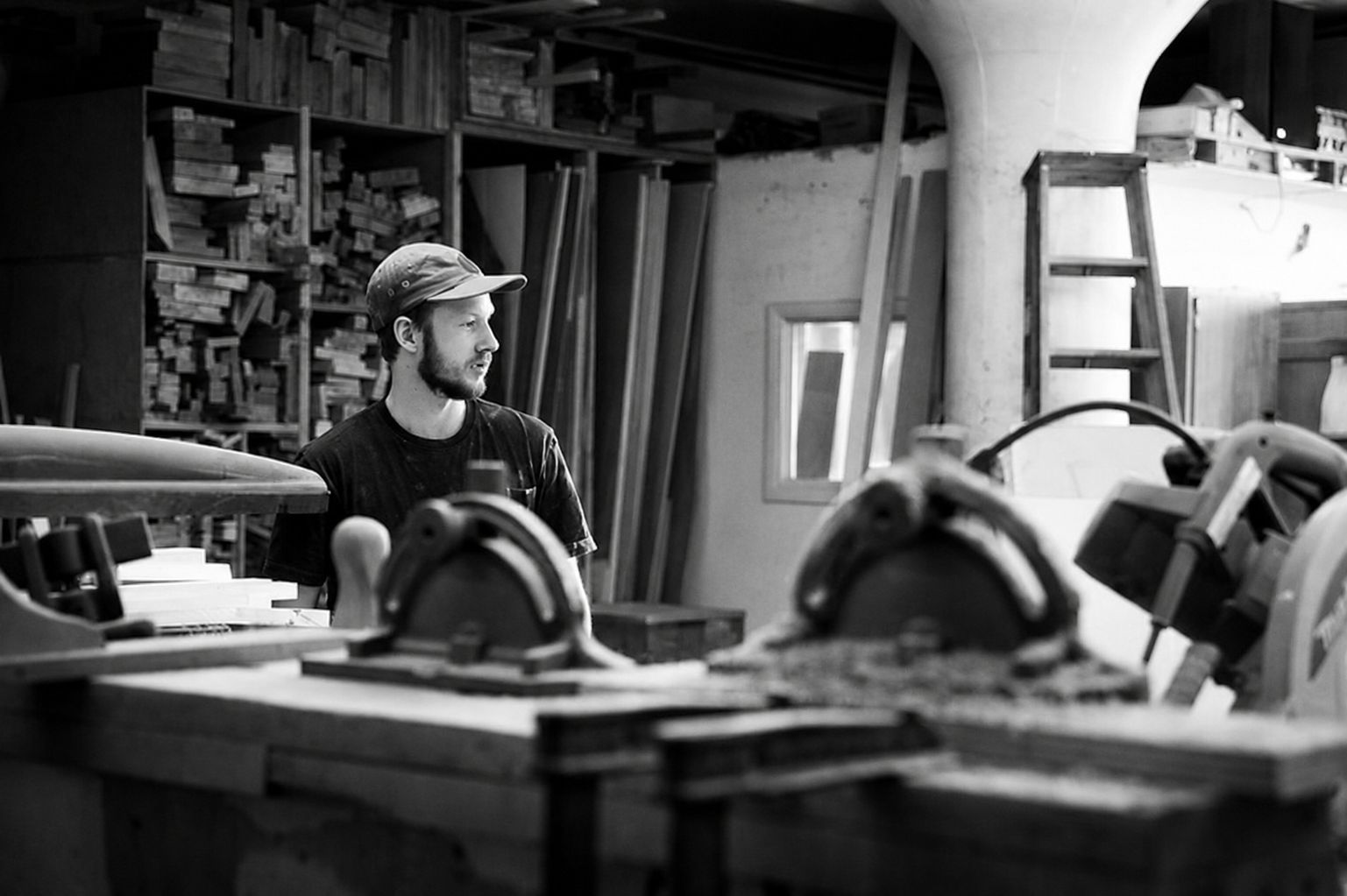 A man with a beard and cap working in a woodworking shop with various tools, wooden planks, and storage shelves around him.