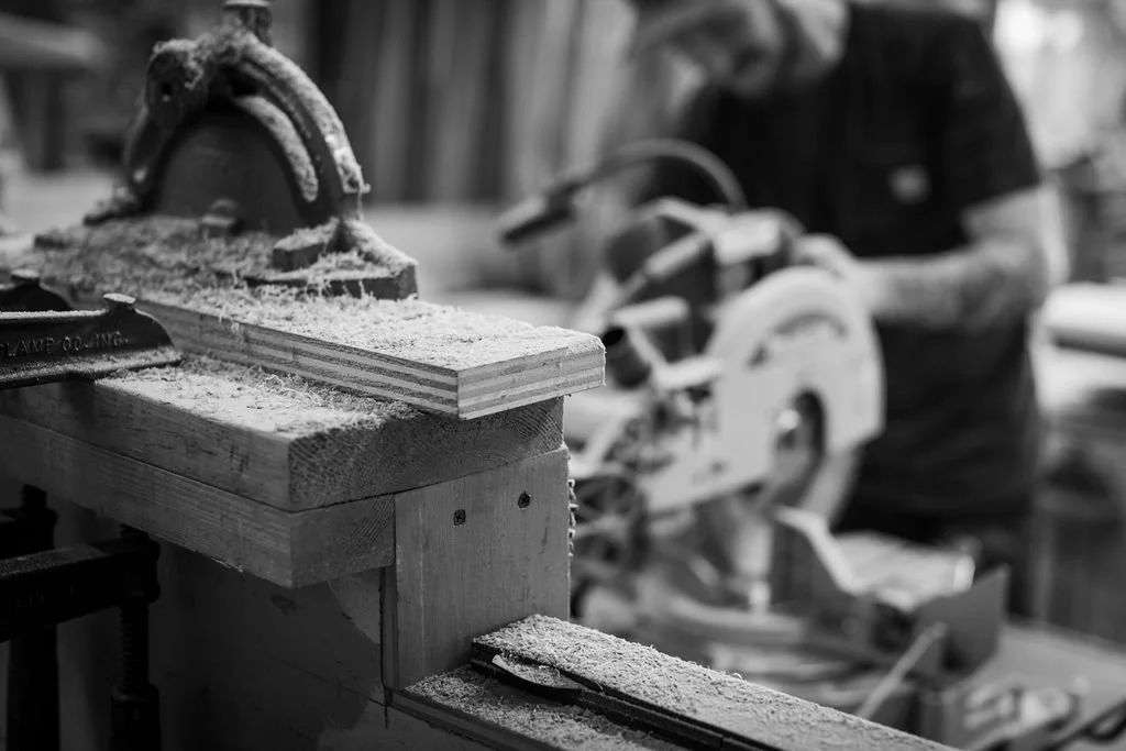 A man working in a woodworking shop, using a circular saw to cut a piece of wood, with sawdust on the workbench.