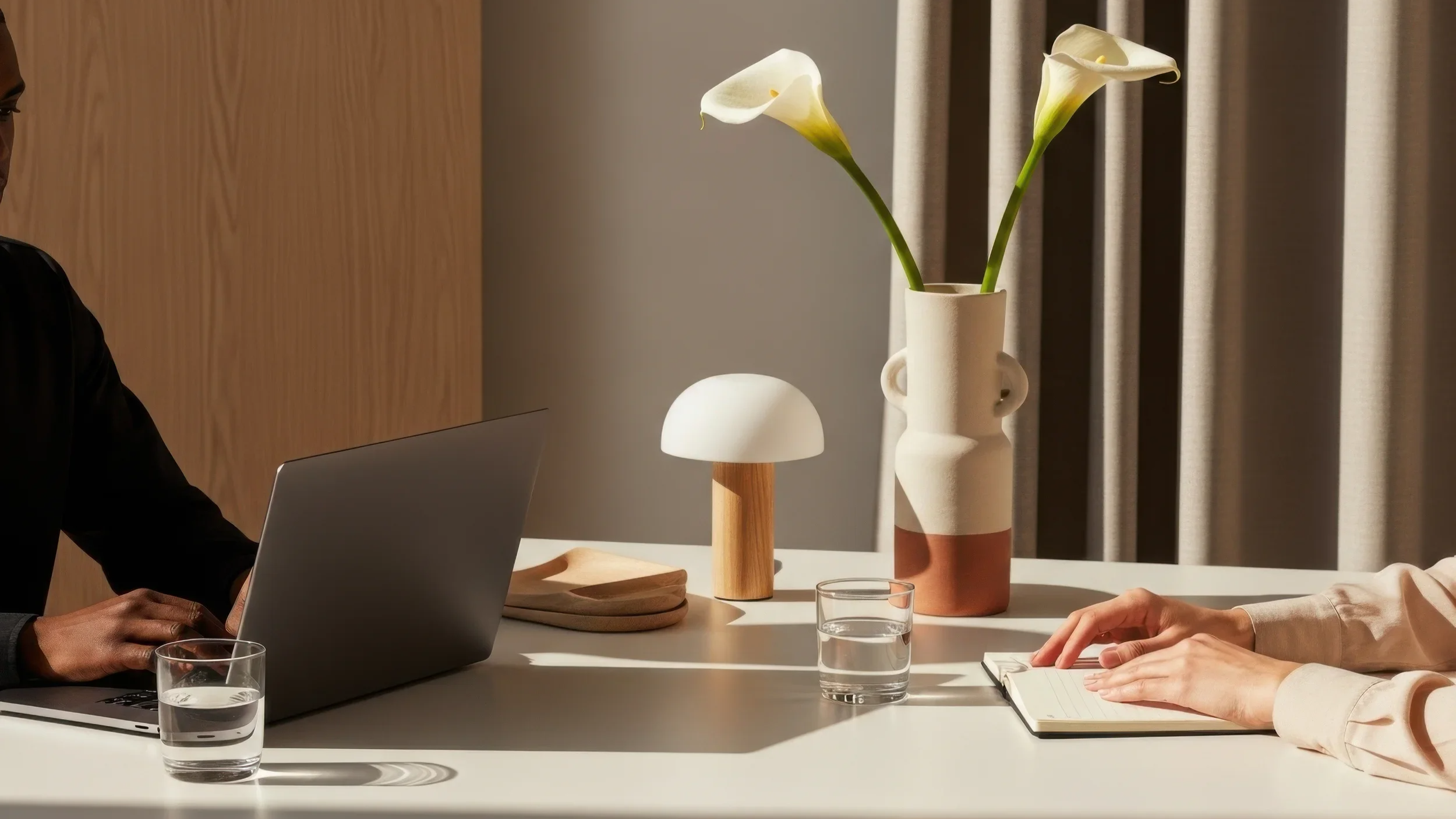 Two people at a meeting table with a laptop, glasses of water, and decorative vases with calla lilies, in a modern office setting.