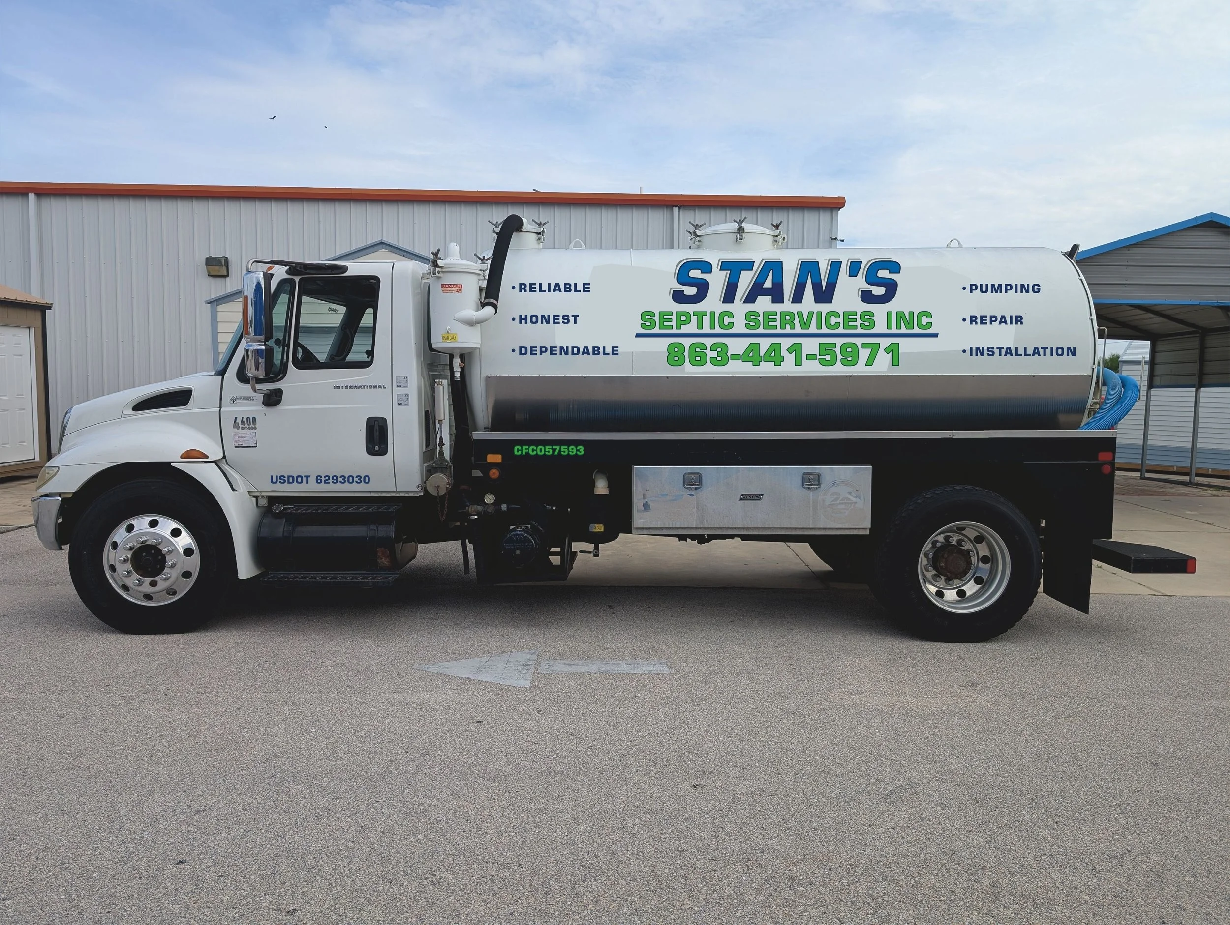 A white vacuum truck parked outdoors near buildings, with signage advertising septic services and contact number.