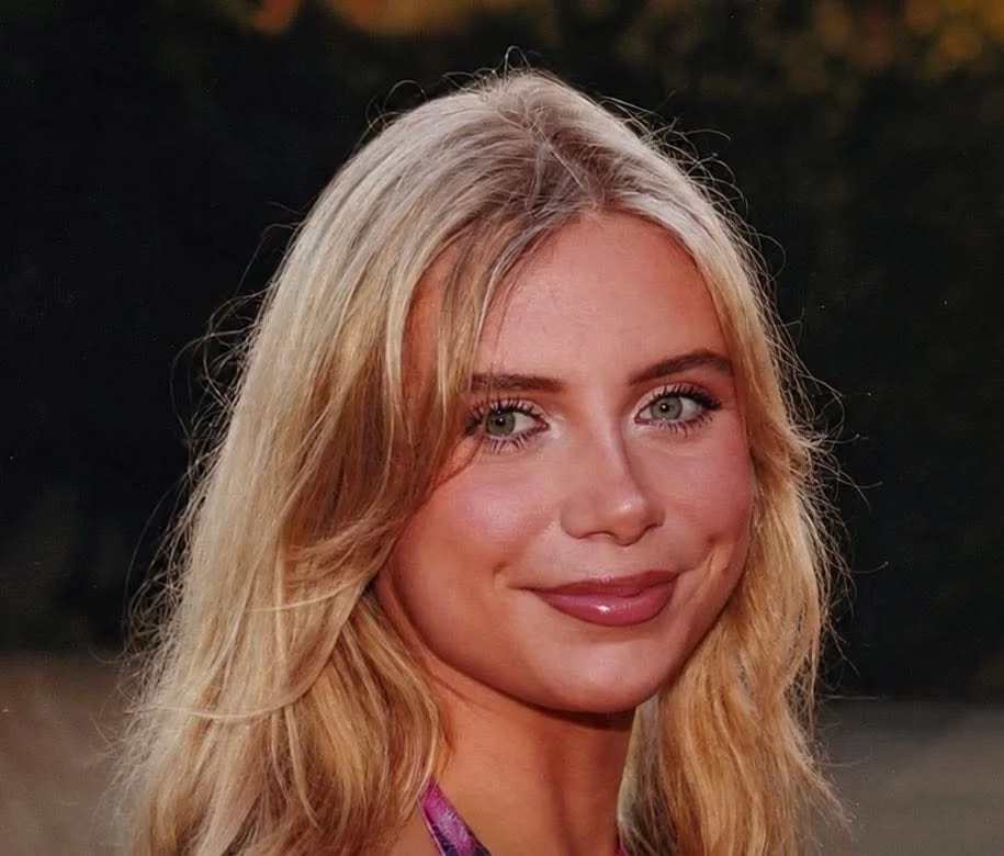 Close-up of a smiling woman with long blonde hair and blue eyes, outdoors at sunset.