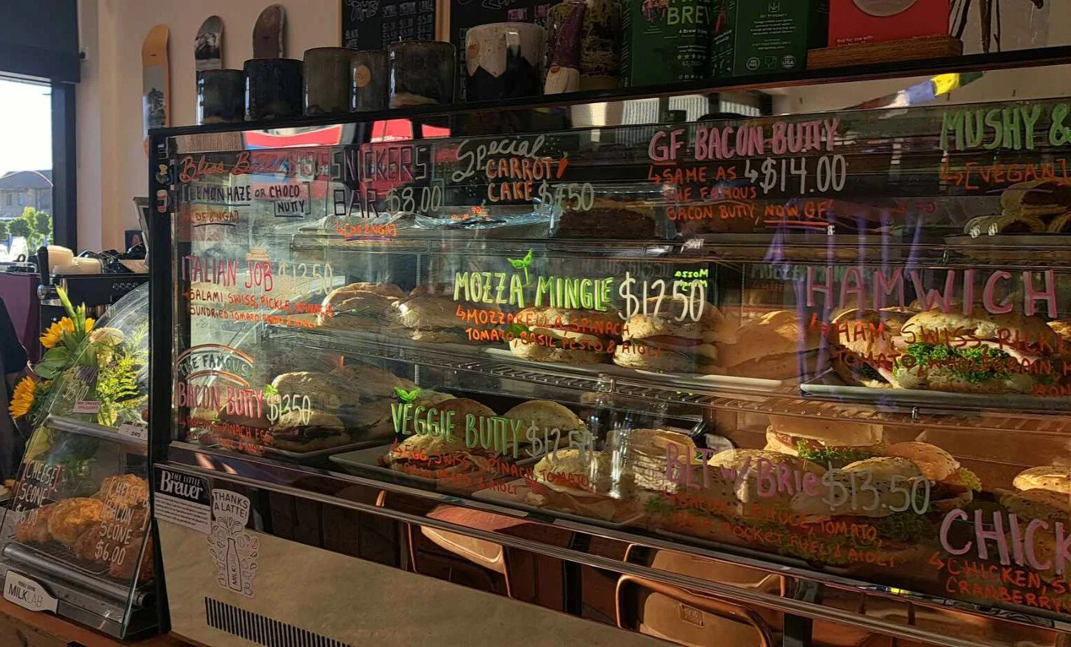 Display case at a café with delicious sandwiches, cakes and bliss balls, decorated with handwritten colorful menu with coffee mugs and espresso coffee machine in background.