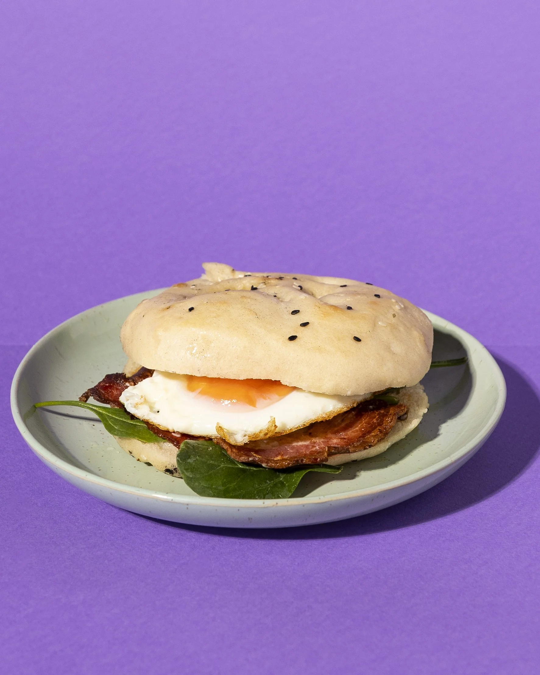 Sandwich with fried egg, bacon, spinach, rocket fuel and aioli on a bun, served on a white plate against a purple background.