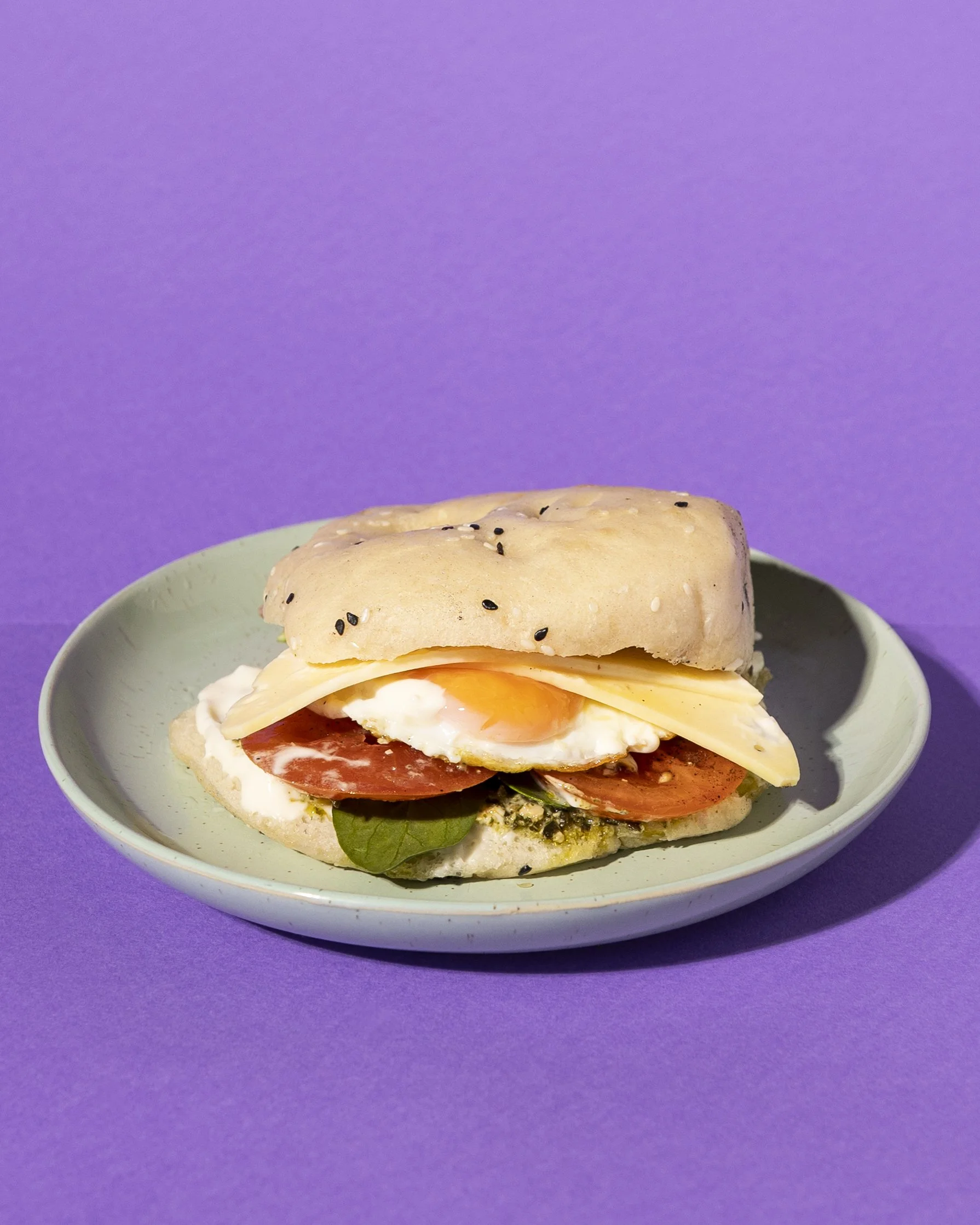 Sandwich on a plate with a purple background, containing an egg, swiss cheese, tomato, spinach, basil pesto and aioli in a bread bun topped with black sesame seeds.