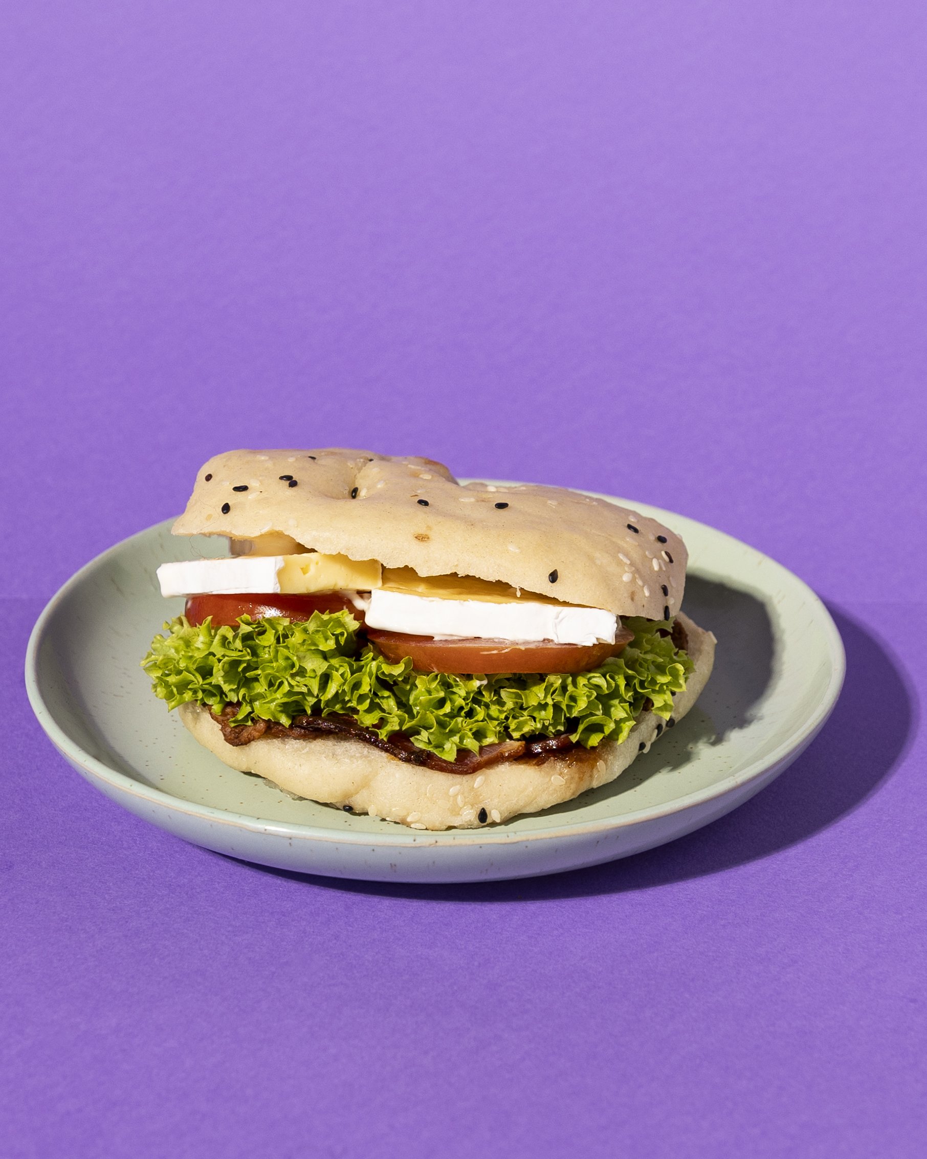 Plate with bread sandwich containing lettuce, tomato, brie cheese, aioli, and bacon against purple background.