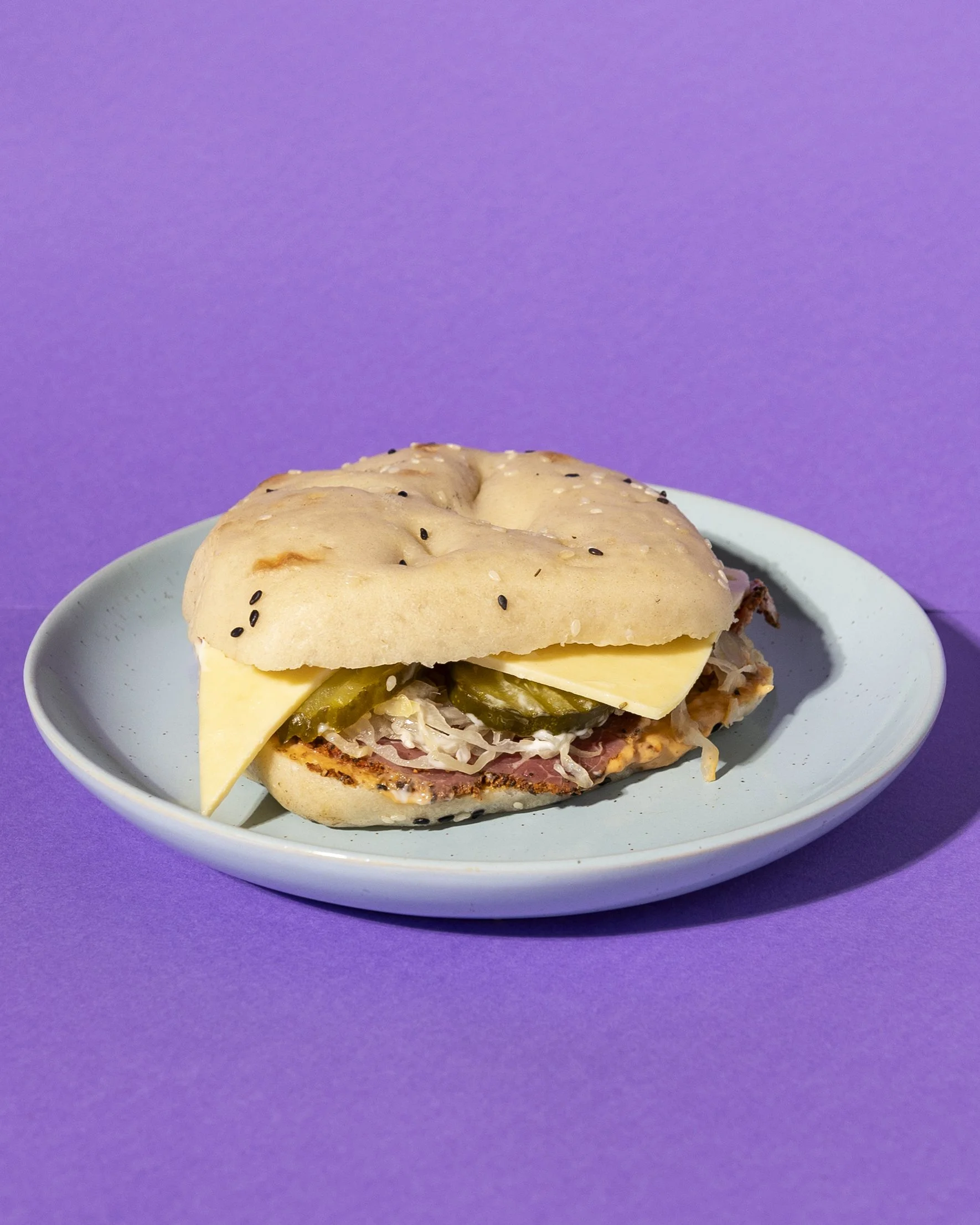 A sandwich on a blue plate with a purple background, containing Swiss cheese, pickles, sauerkraut cabbage, and pastrami with Hoot sauce and aioli, in a bread roll topped with sesame seeds.
