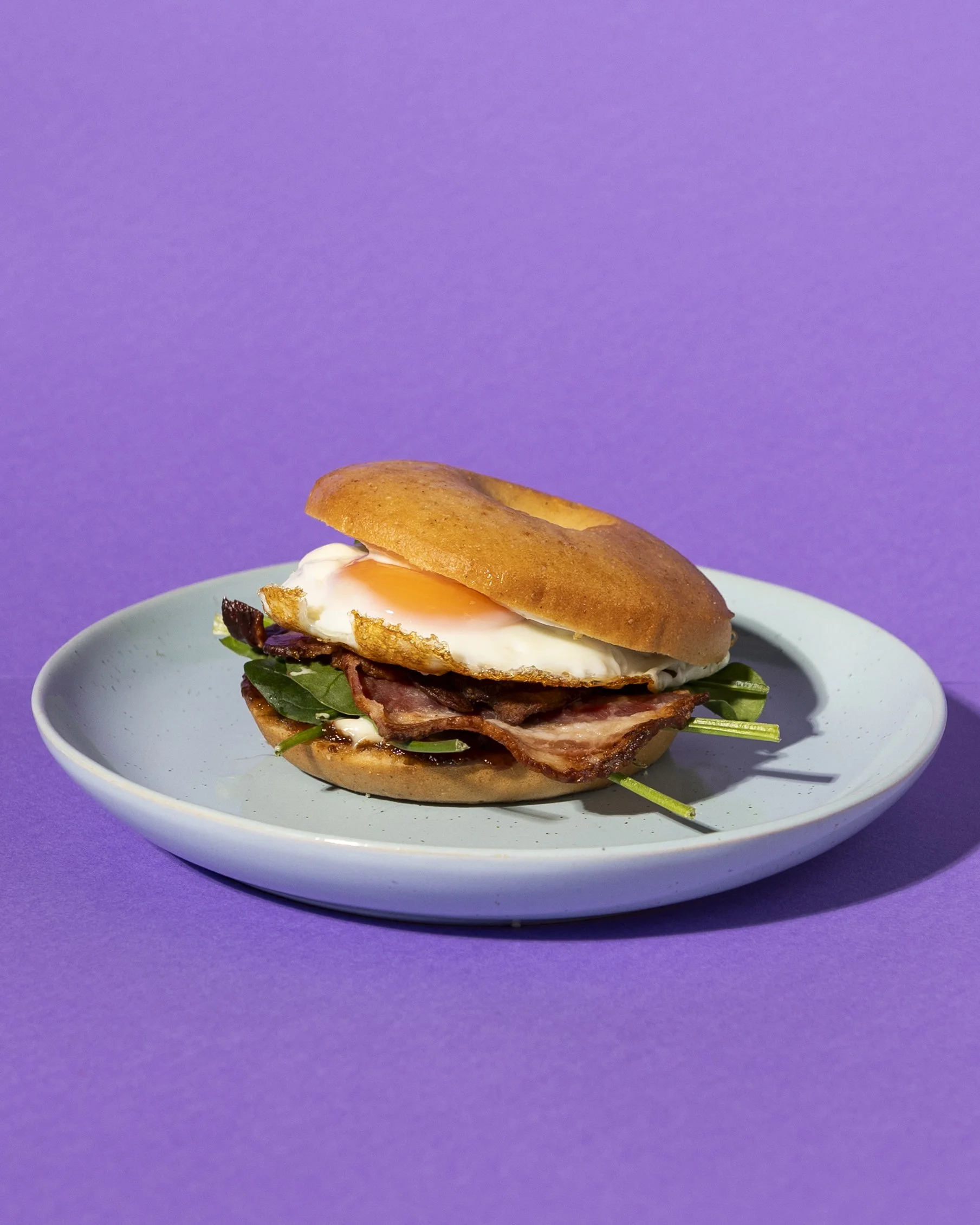 A gluten-free sandwich on a blue plate with bacon, spinach, a fried egg, and a gluten free bagel, with a purple background.