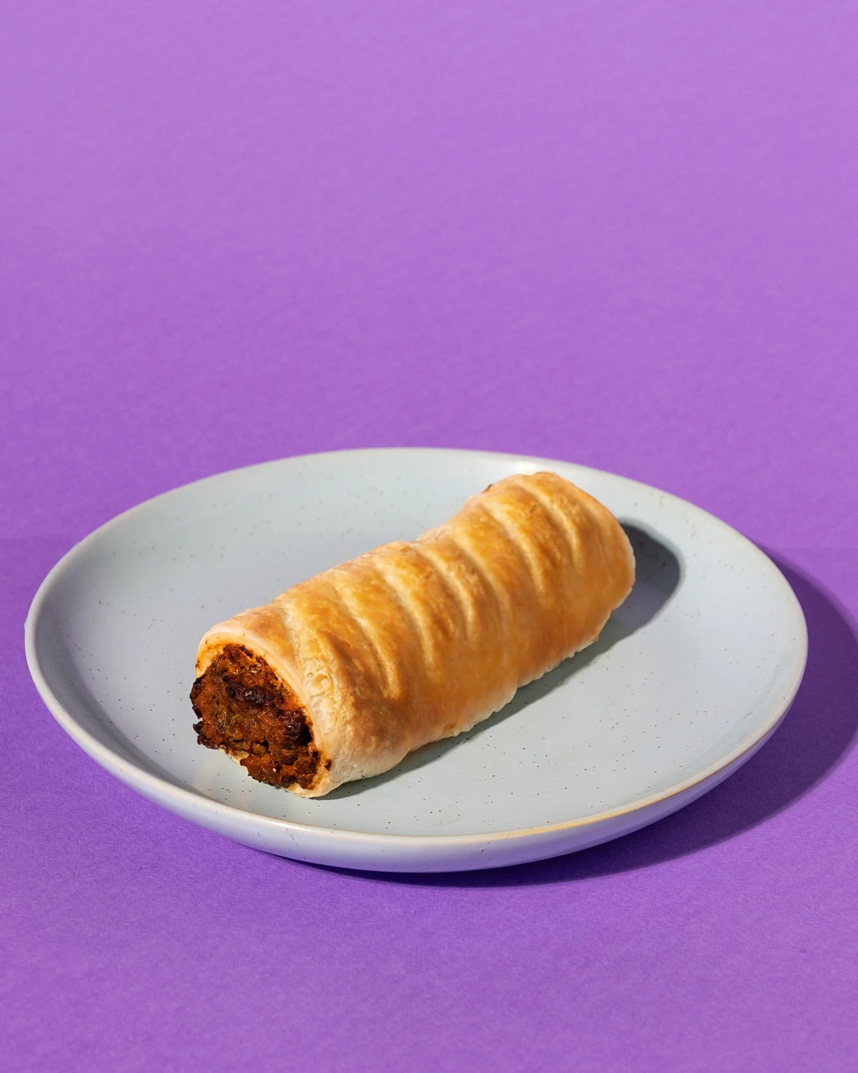 A pastry with a golden, flaky crust on a blue plate, with a mushroom and black bean filling inside, set against a purple background.