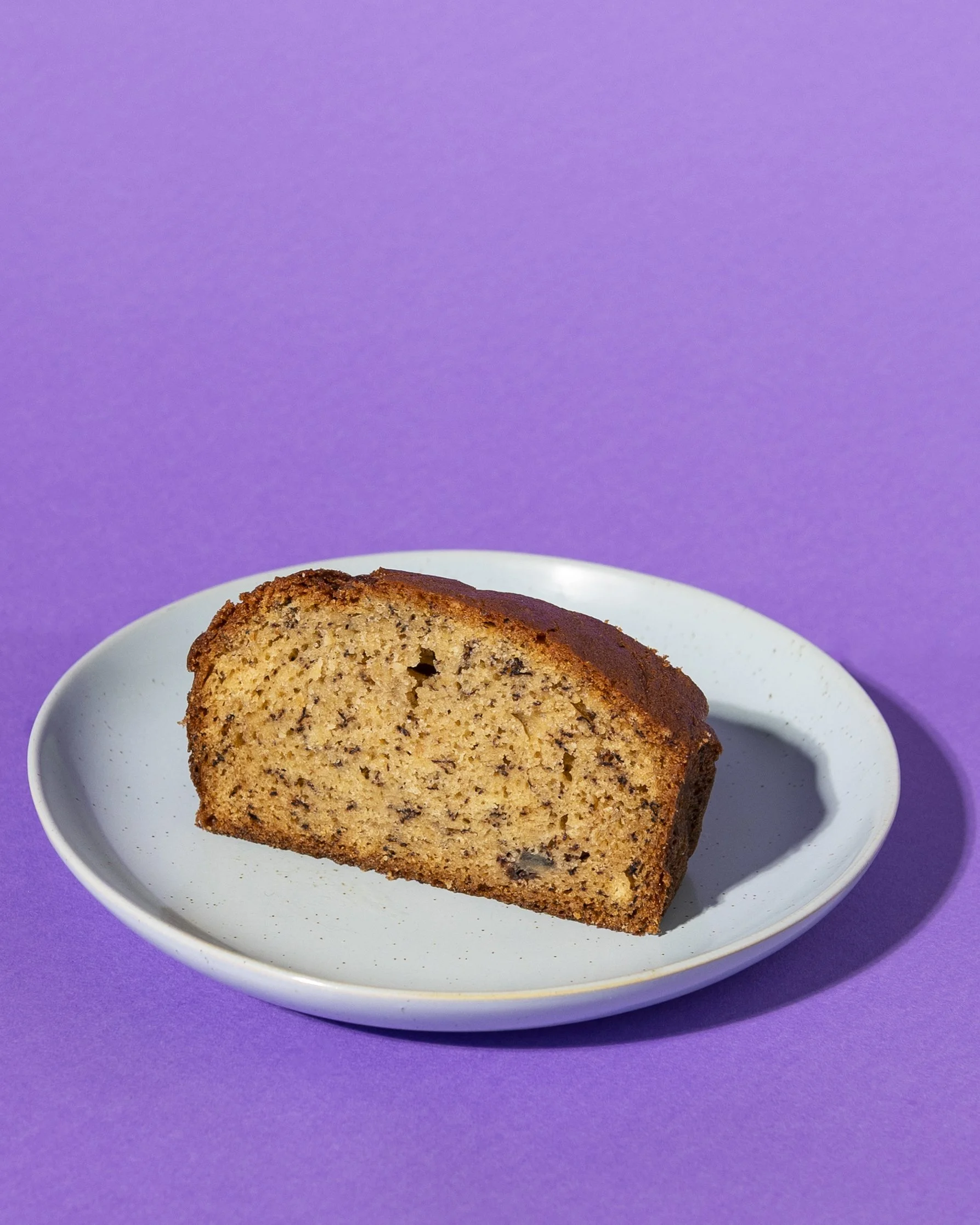 Slice of banana bread on light blue plate against purple background. Served great with fresh coffee