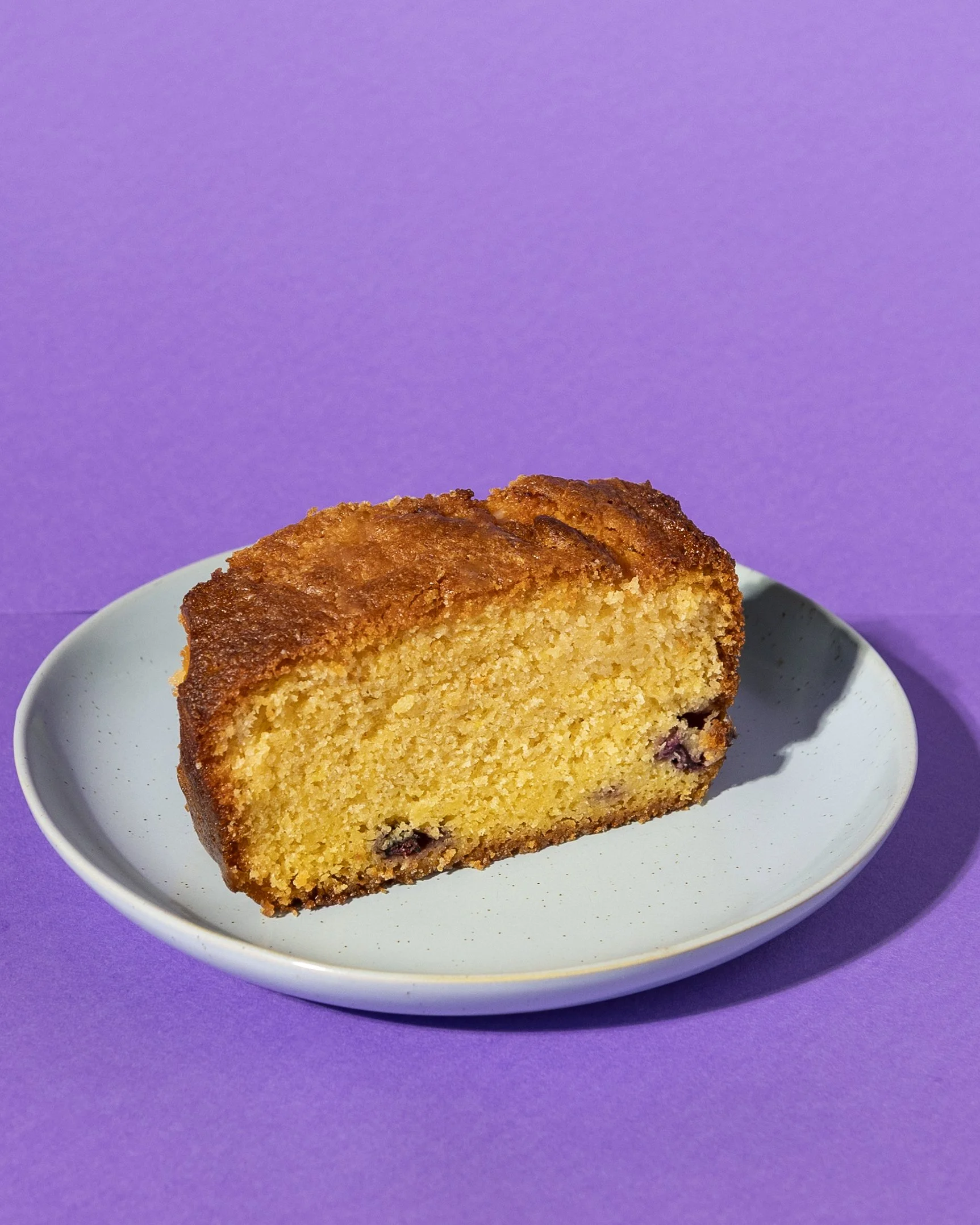 Slice of lemon drizzle cake on a light blue plate against a purple background. Goes well with a freshly brewed coffee.