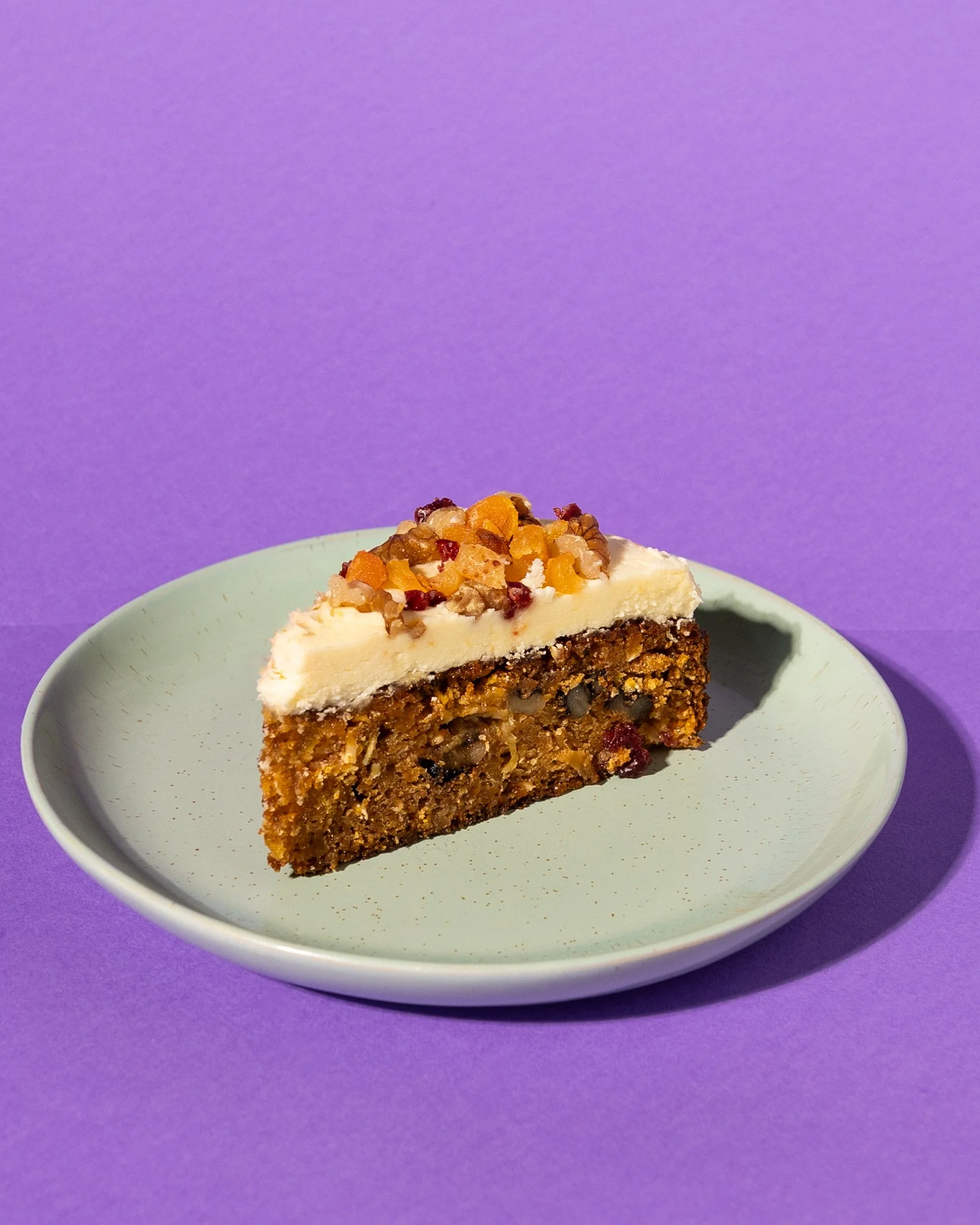 Slice of carrot cake with lemon butter cream icing, topped with chopped nuts and dried pineapple, cranberry and walnuts on a light green plate against a purple background. Goes great with freshly brewed espresso coffee