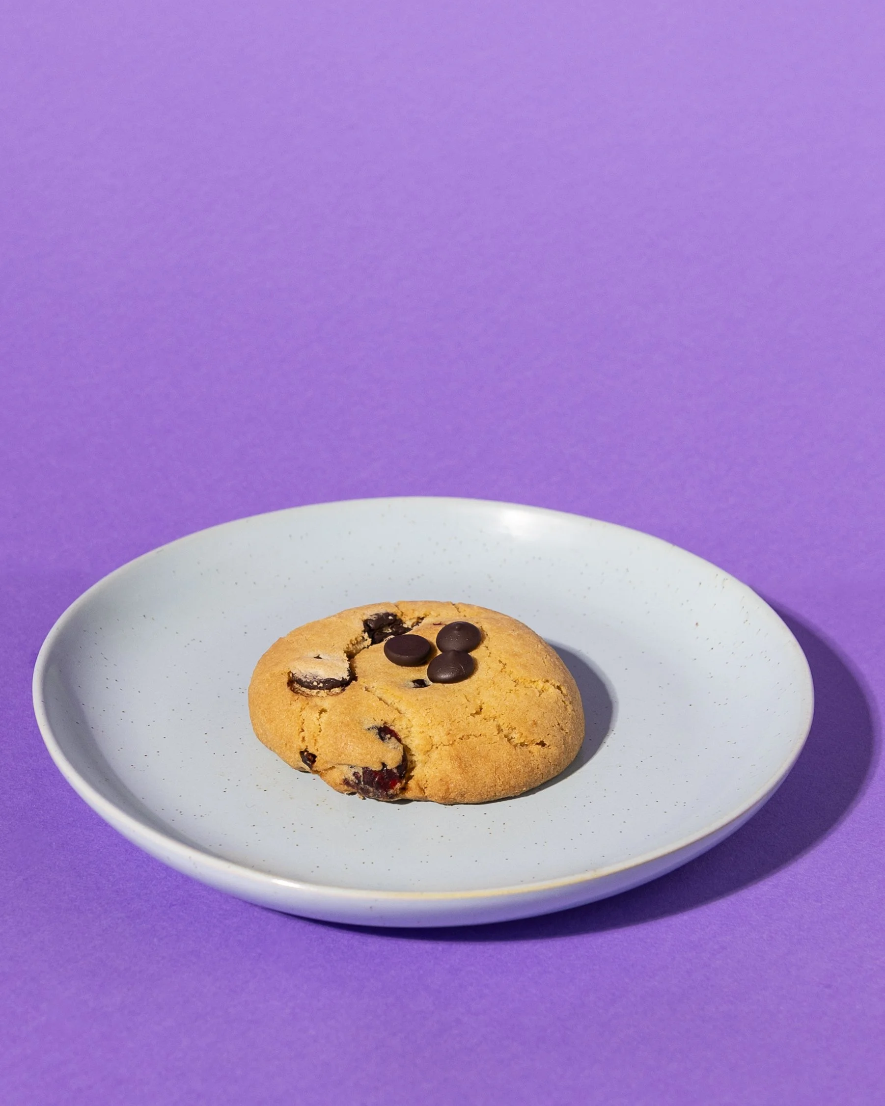 A chocolate chip cookie on a light blue plate against a purple background. Rotating cookie flavours. Served best with in-house roasted coffee beans.