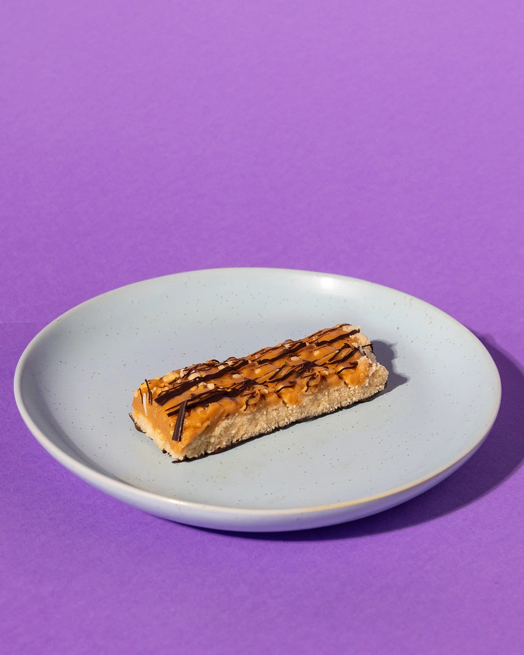A slice of dessert with a crumbly base, topped with caramel, chopped nuts, and drizzled with chocolate, on a blue plate against a purple background. Served best with a fresh coffee.