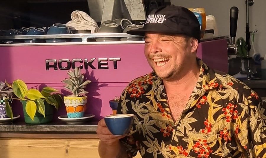 A man wearing a floral shirt and a black cap with white text, smiling and holding a blue and white coffee cup, standing in front of a pink coffee machine labeled "Rocket," with potted plants nearby.