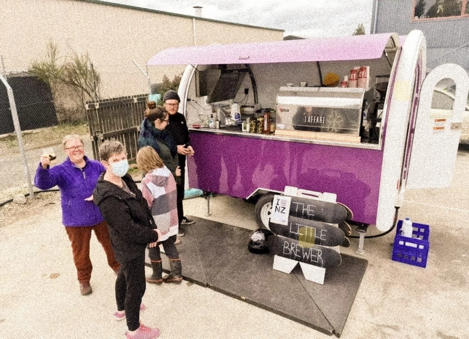 A purple mobile coffee cart with a pink canopy, serving coffee and beverages, is set up outdoors. Four people, one wearing a face mask, are standing in line in front of the cart, with one person excitedly raising a glass. A blackboard sign says 'NZ THE LITTLE BREWER'.