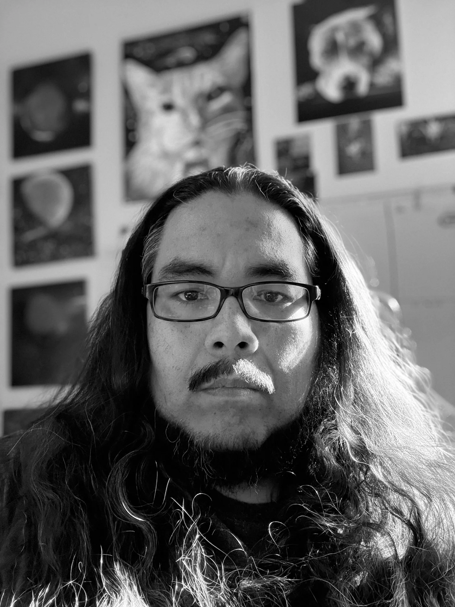 Black and white photo of a person with long curly hair, wearing glasses, with framed space-themed artwork on the wall behind them.