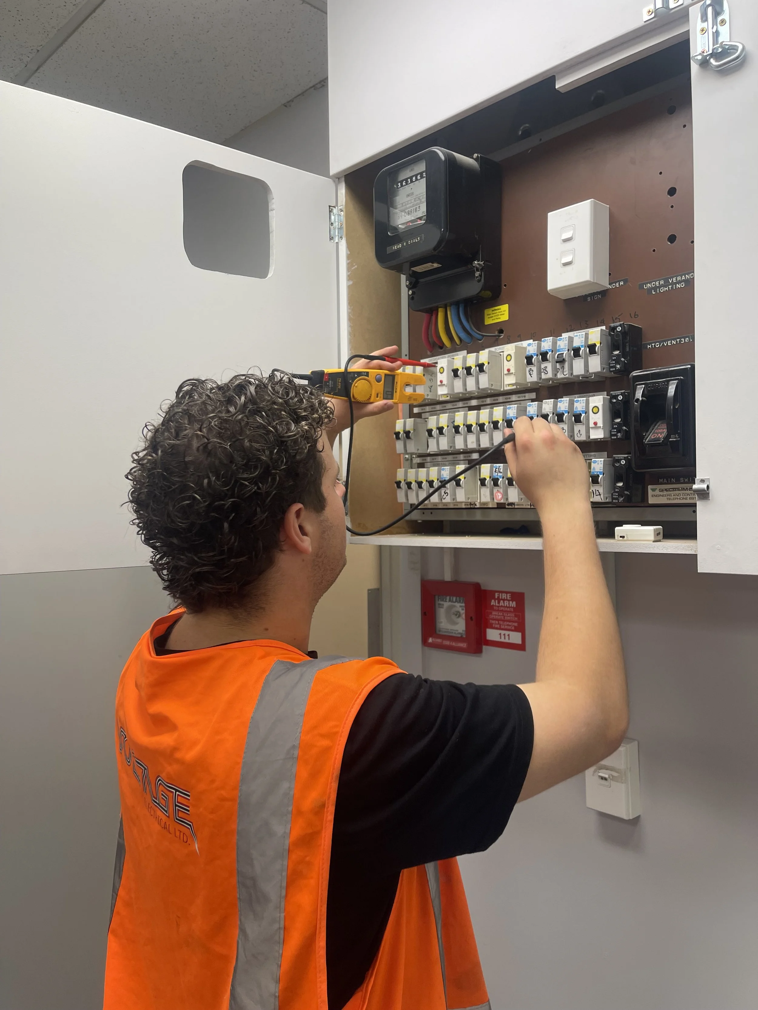 A technician wearing an orange safety vest using a multimeter on an electrical panel.