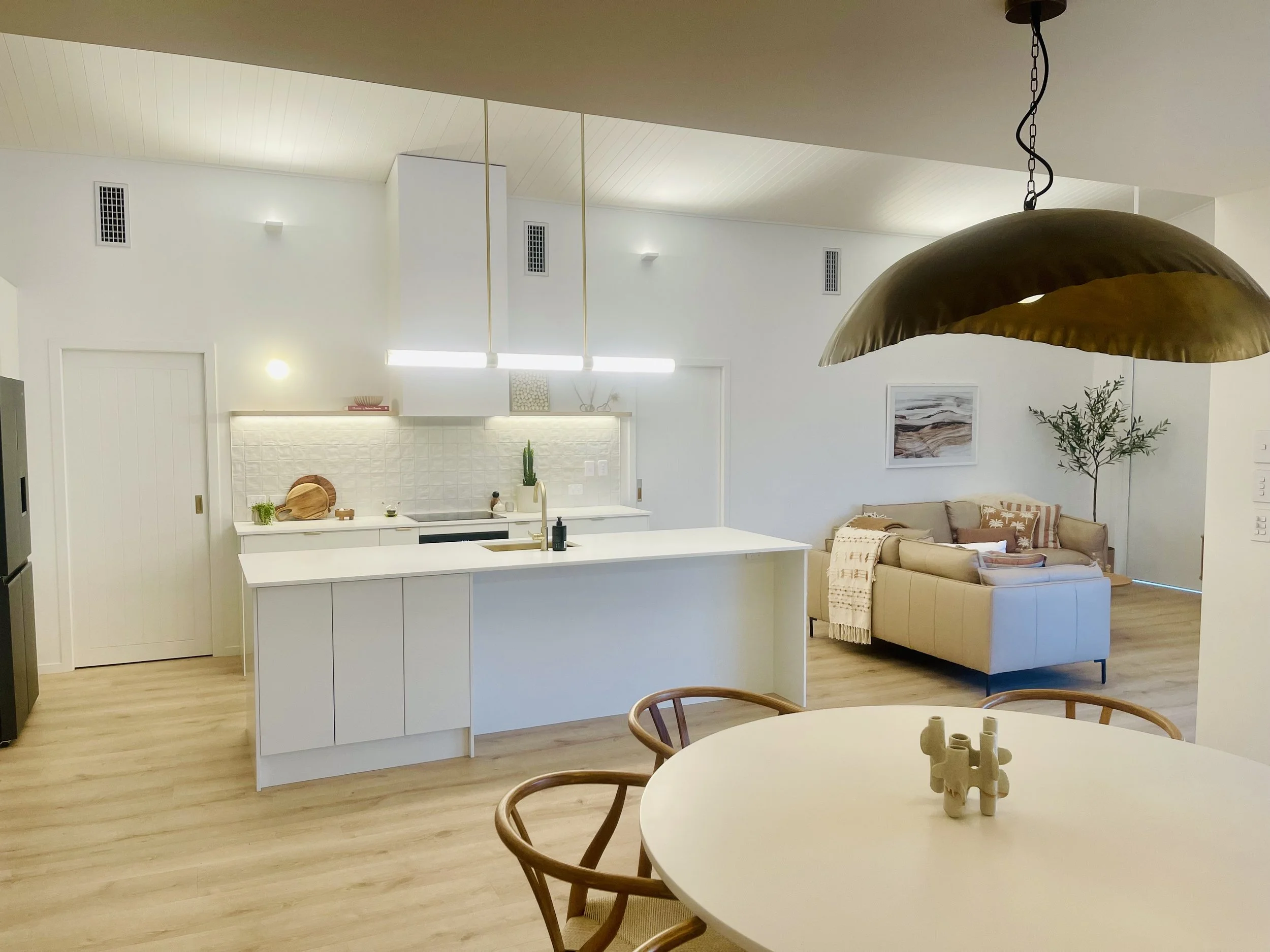 Open-concept kitchen and living room space with white walls, light wood flooring, a white kitchen island, a beige sofa with patterned pillows, a dining table with wooden chairs, a large gold pendant light, and wall art.