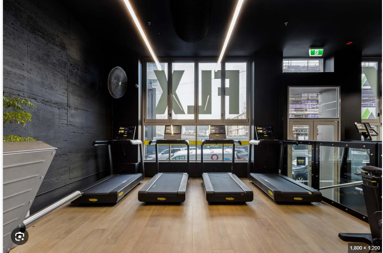 Indoor gym with four treadmills facing large windows with city view, modern black and wood interior, and gym equipment.
