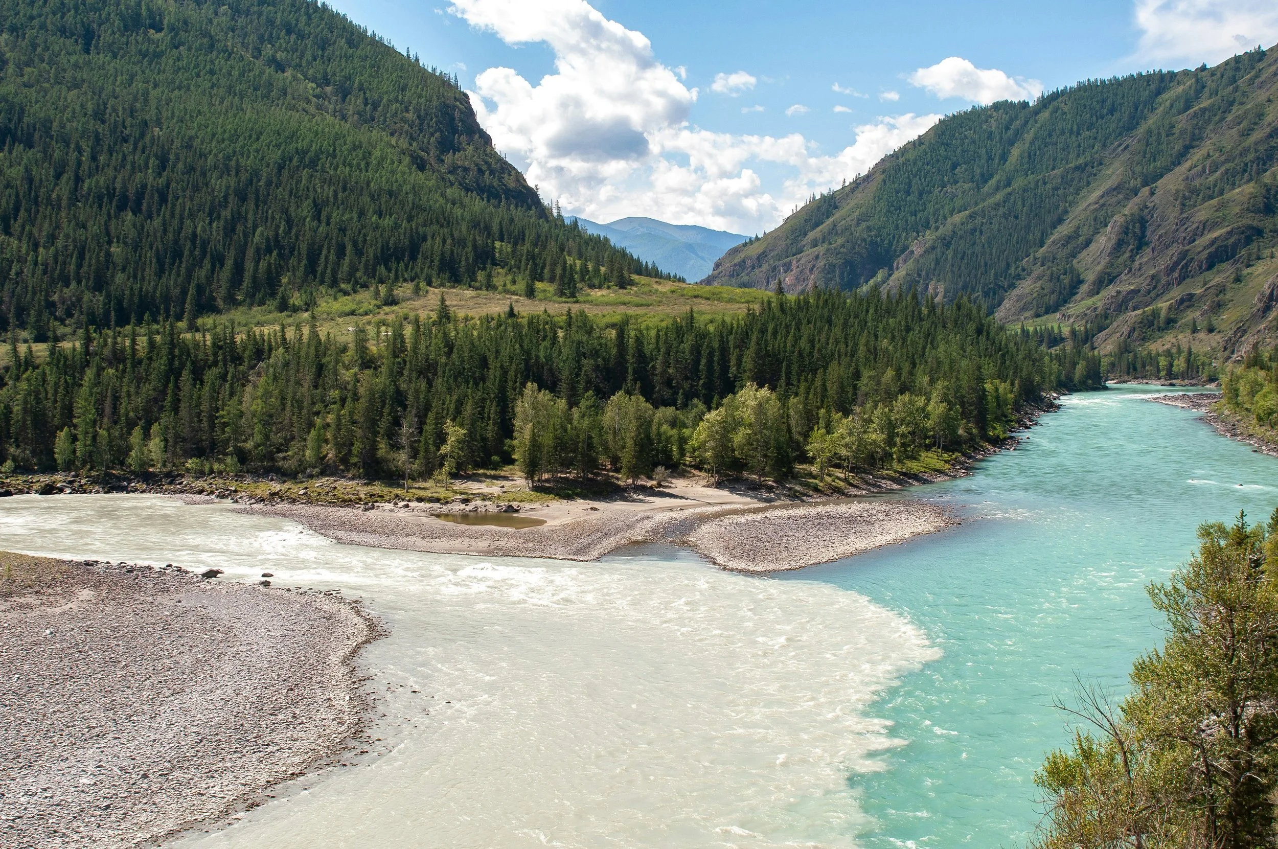A river flowing through a lush green mountainous landscape with scattered trees, blue sky, and white clouds.