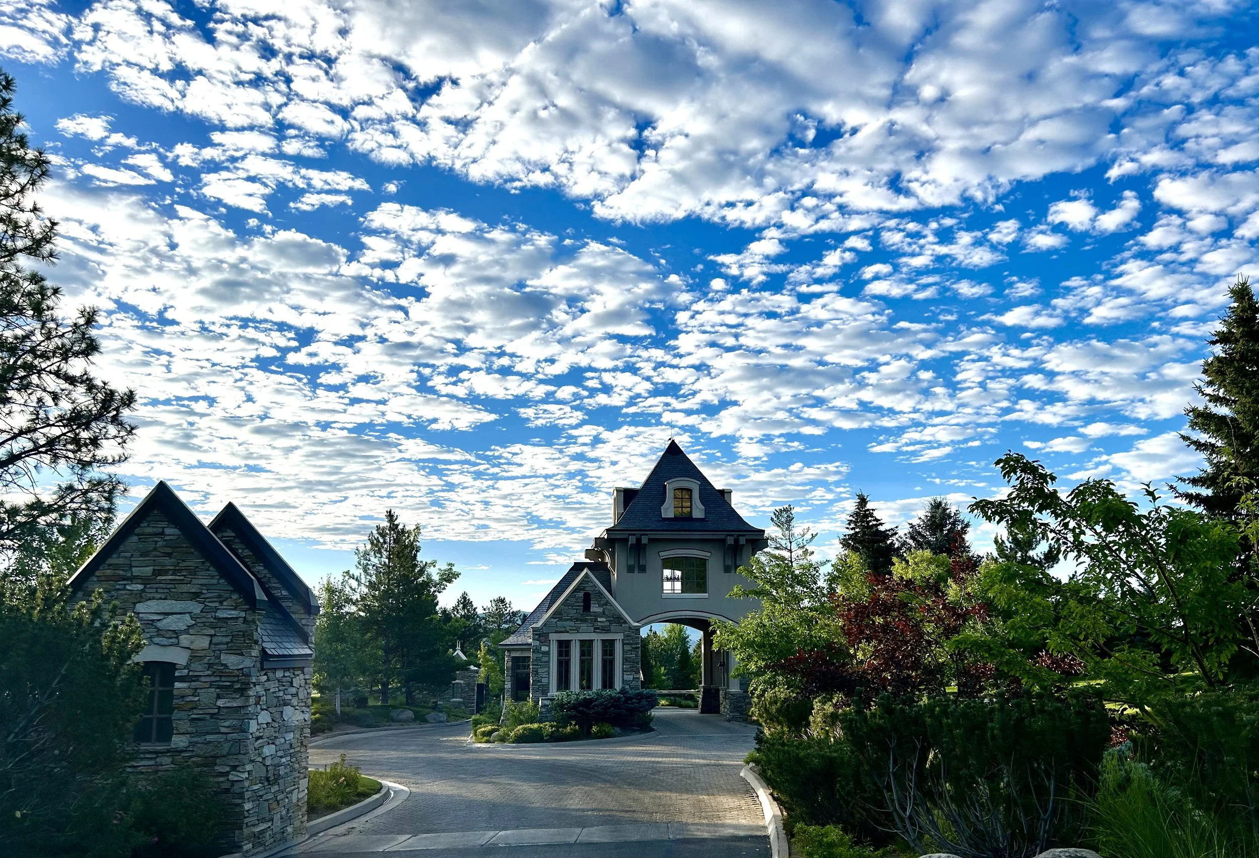 View of the Montreux gatehouse with a pointed turret roof, stone accents, surrounded by lush greenery and trees, under a bright blue sky with scattered white clouds.