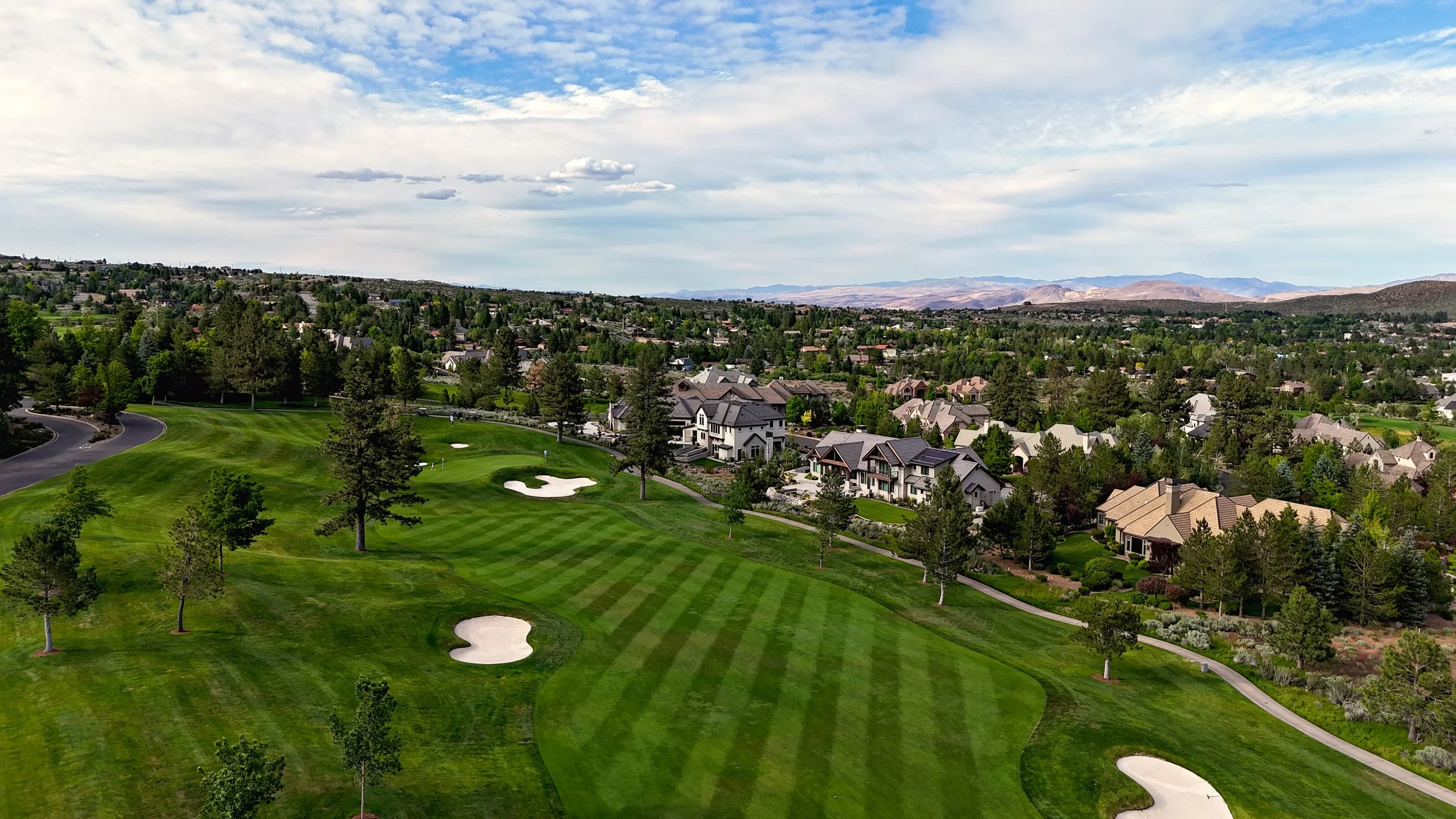 A golf course with lush green grass, sand bunkers, and scattered trees in the foreground. Residential houses and buildings are visible beyond the course with a mountainous landscape in the background. The sky is partly cloudy.