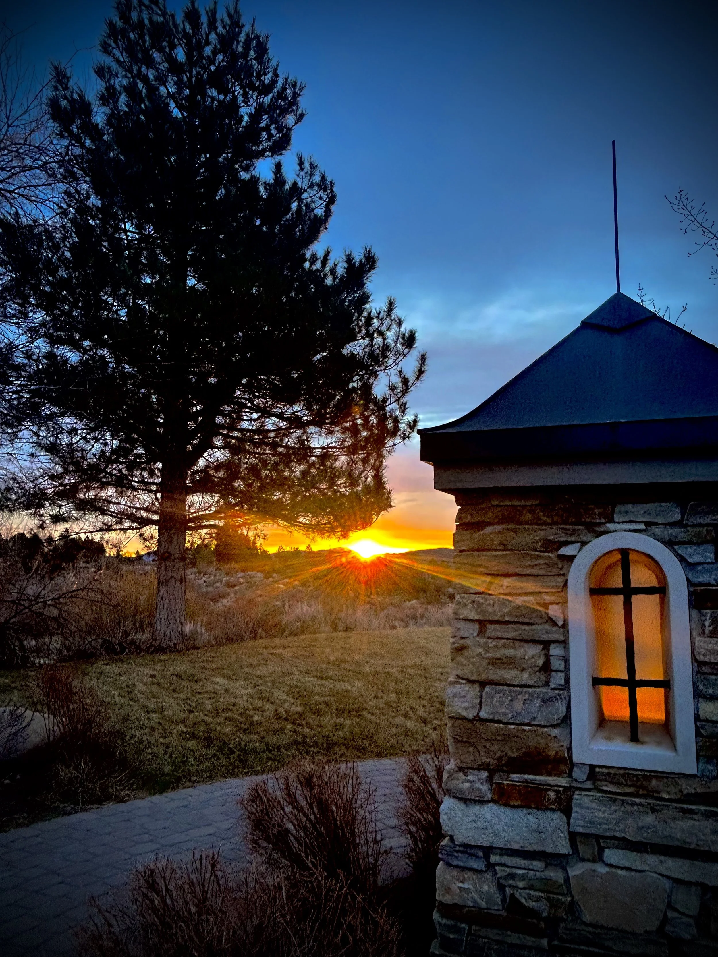 Photo of a Montreux entry landscape during sunset with a large tree on the left, a stone gate with a window on the right, and a colorful sky transitioning from blue to orange in the background.