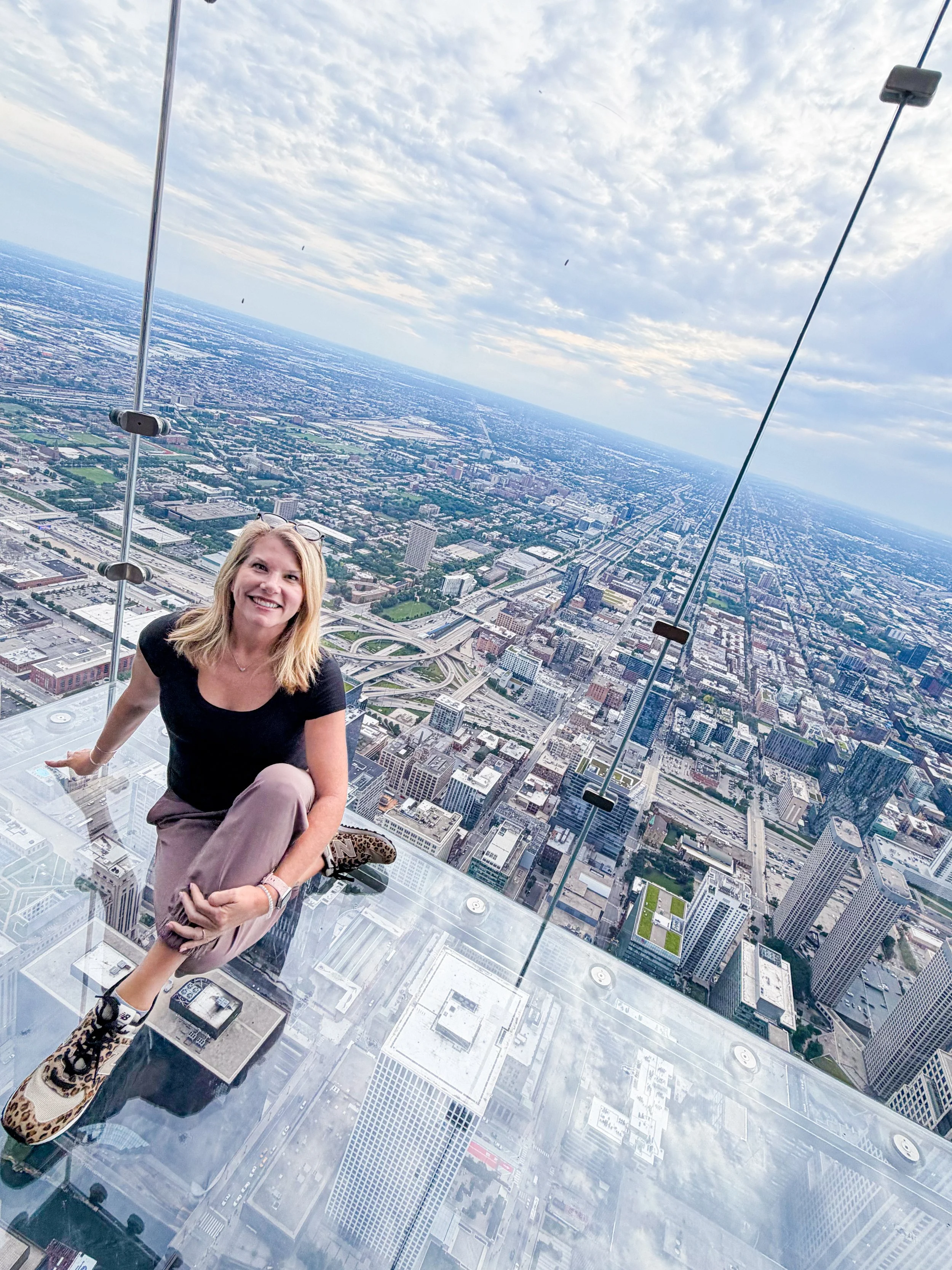 Smiling traveler sitting on a glass observation ledge high above a city skyline