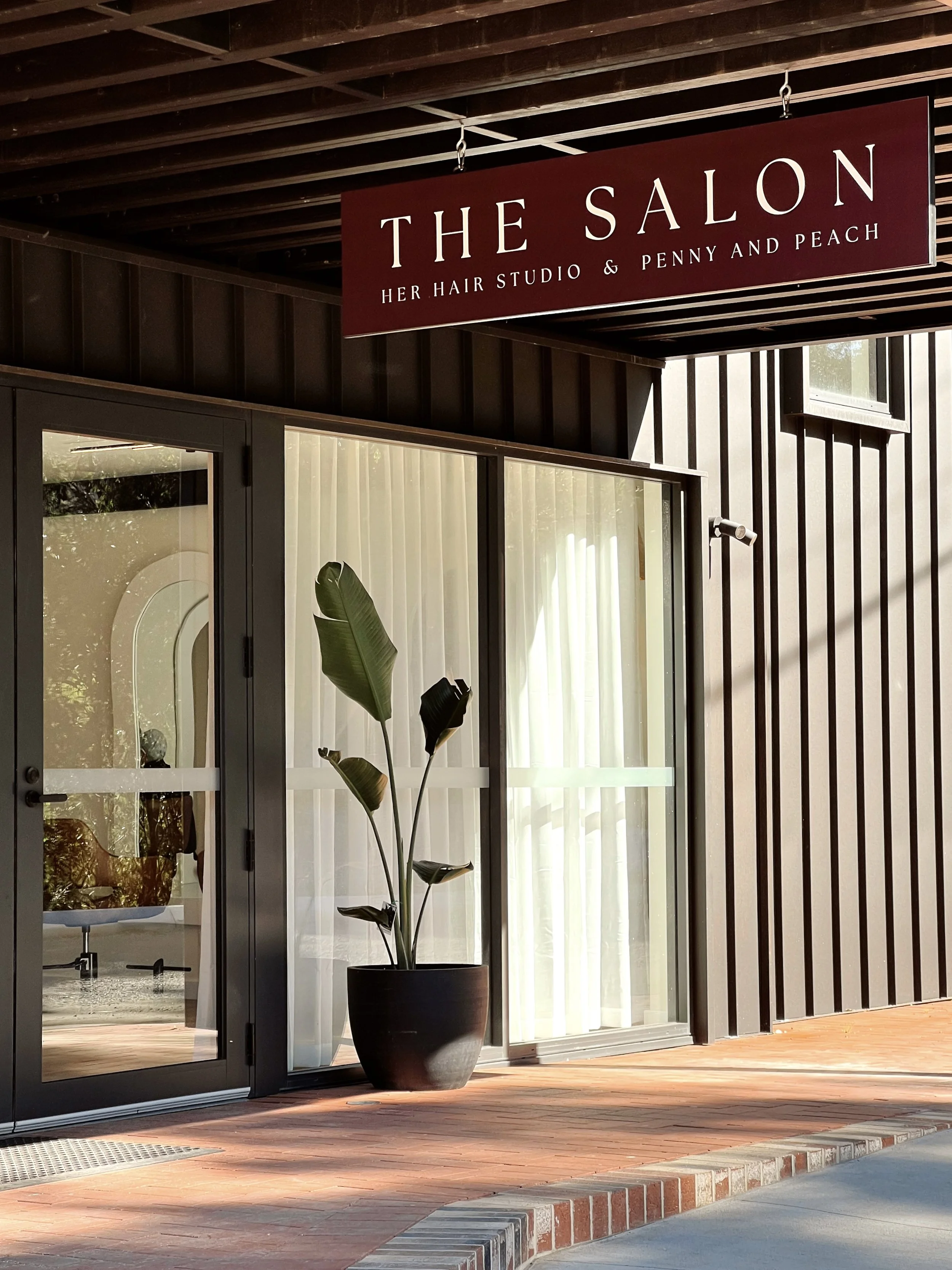 Storefront with a sign reading 'THE SALON HER HAIR STUDIO & PENNY AND PEACH', large glass windows, a potted plant, and a brick sidewalk.