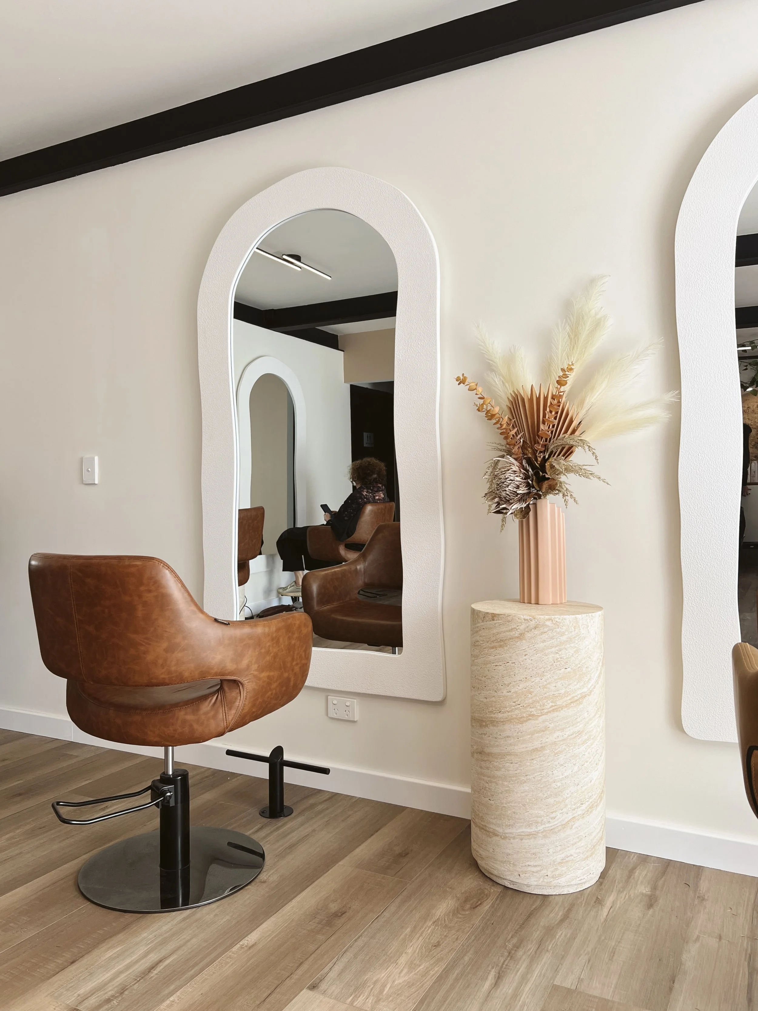 Modern salon interior with a brown swivel chair, large decorative vase with dried flowers, and a white textured mirror on a beige wall.